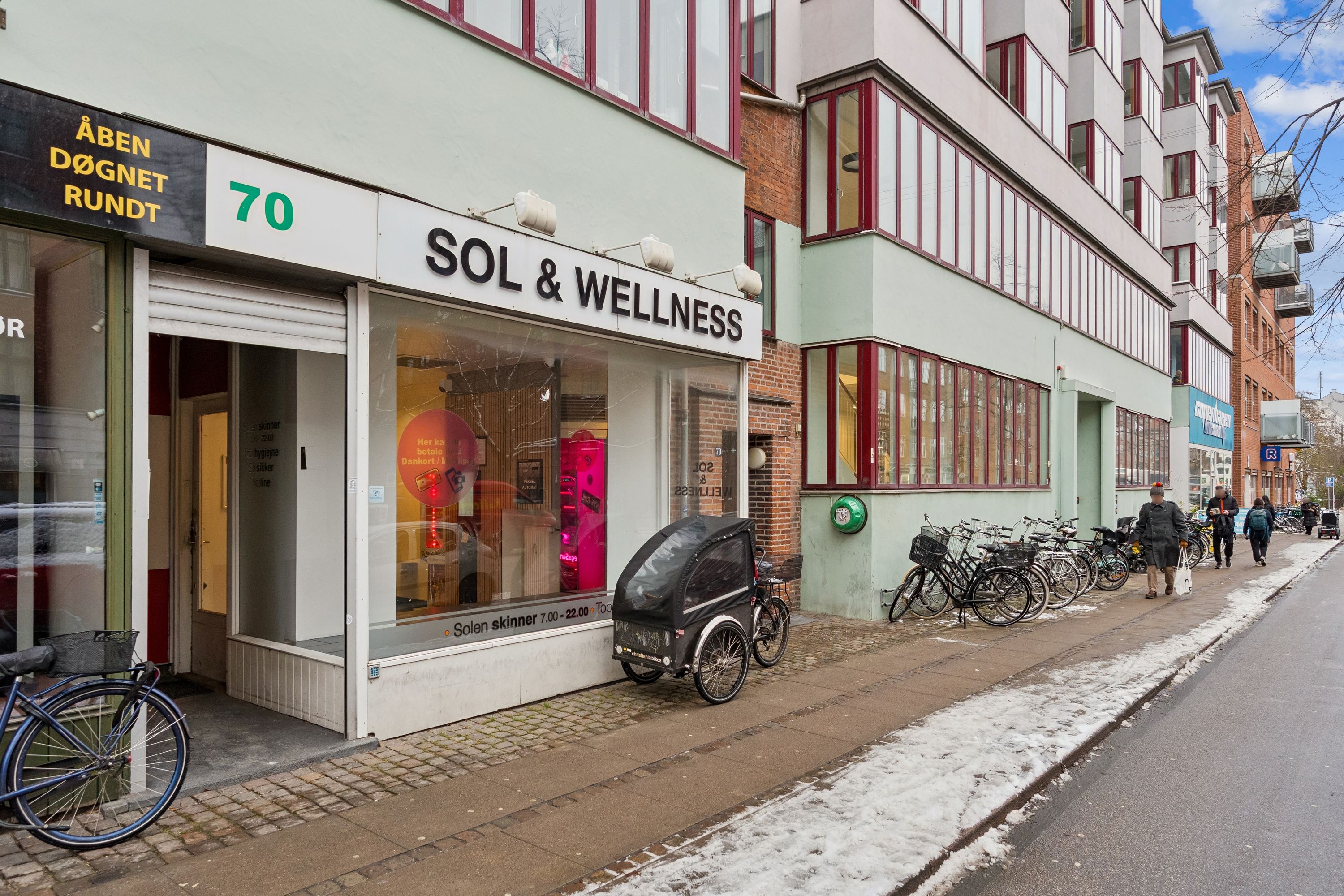 Sol & Wellness butik med stående sollampe og tre cykler udenfor på Jagtvej 70 i København. Adgangsvej og cykelparkering med folk og sne på siderne