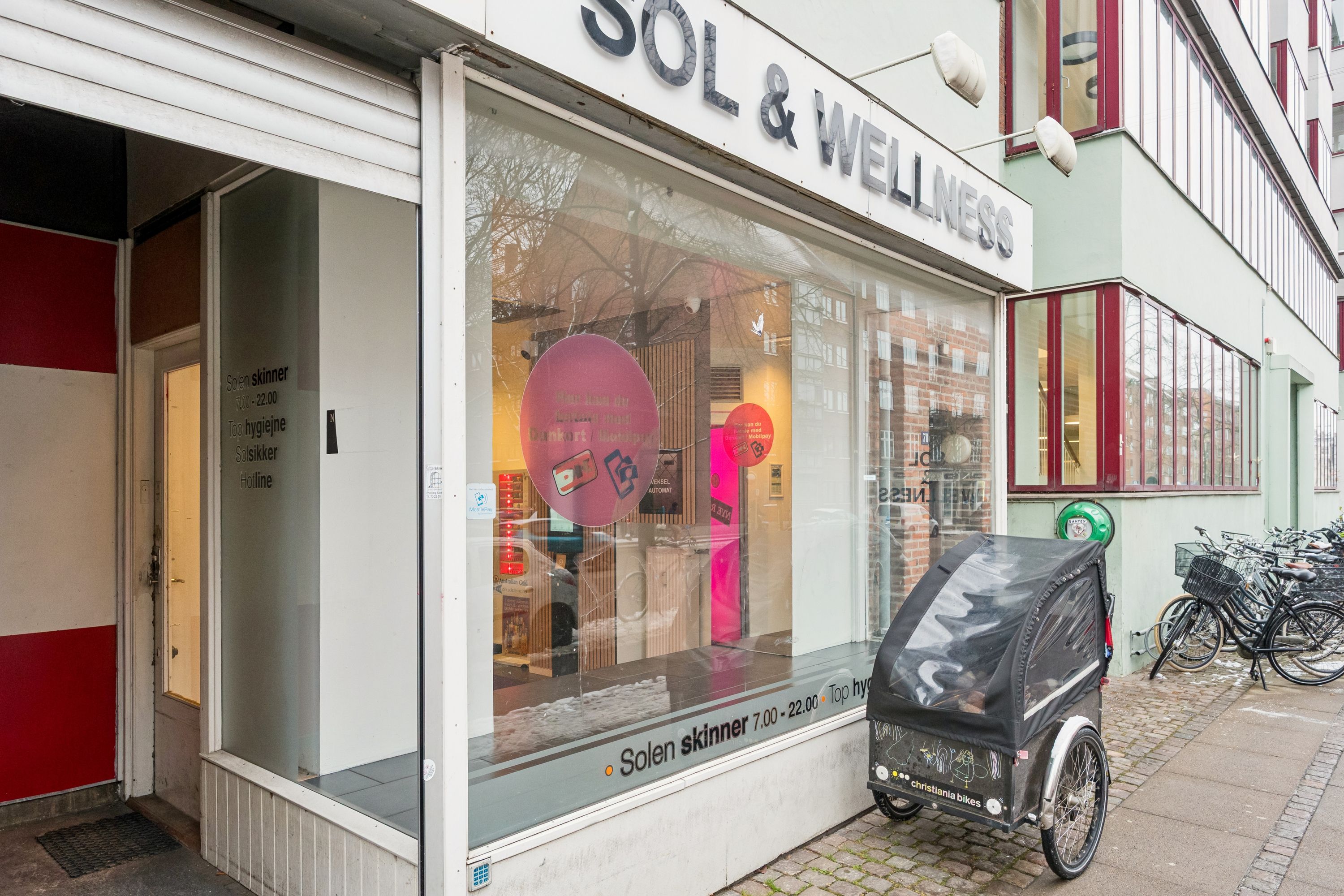 En sol- og wellness-butik med glasruder og stående skilte med åbningstider og Sol-søjler, uden for er en cykeltrailer og cykler parkeret