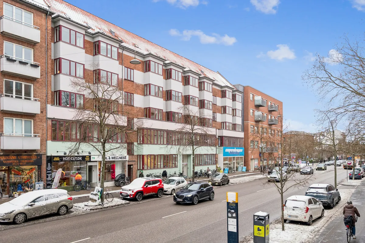 Butik til leje på Jagtvej 70, st. th, 2200 København N. Bygning med røde mursten og hvide vindueskarme. Butikker med skilte som Hot-Cut.dk og OL & Wellness. Både biler og cykler står parkeret.