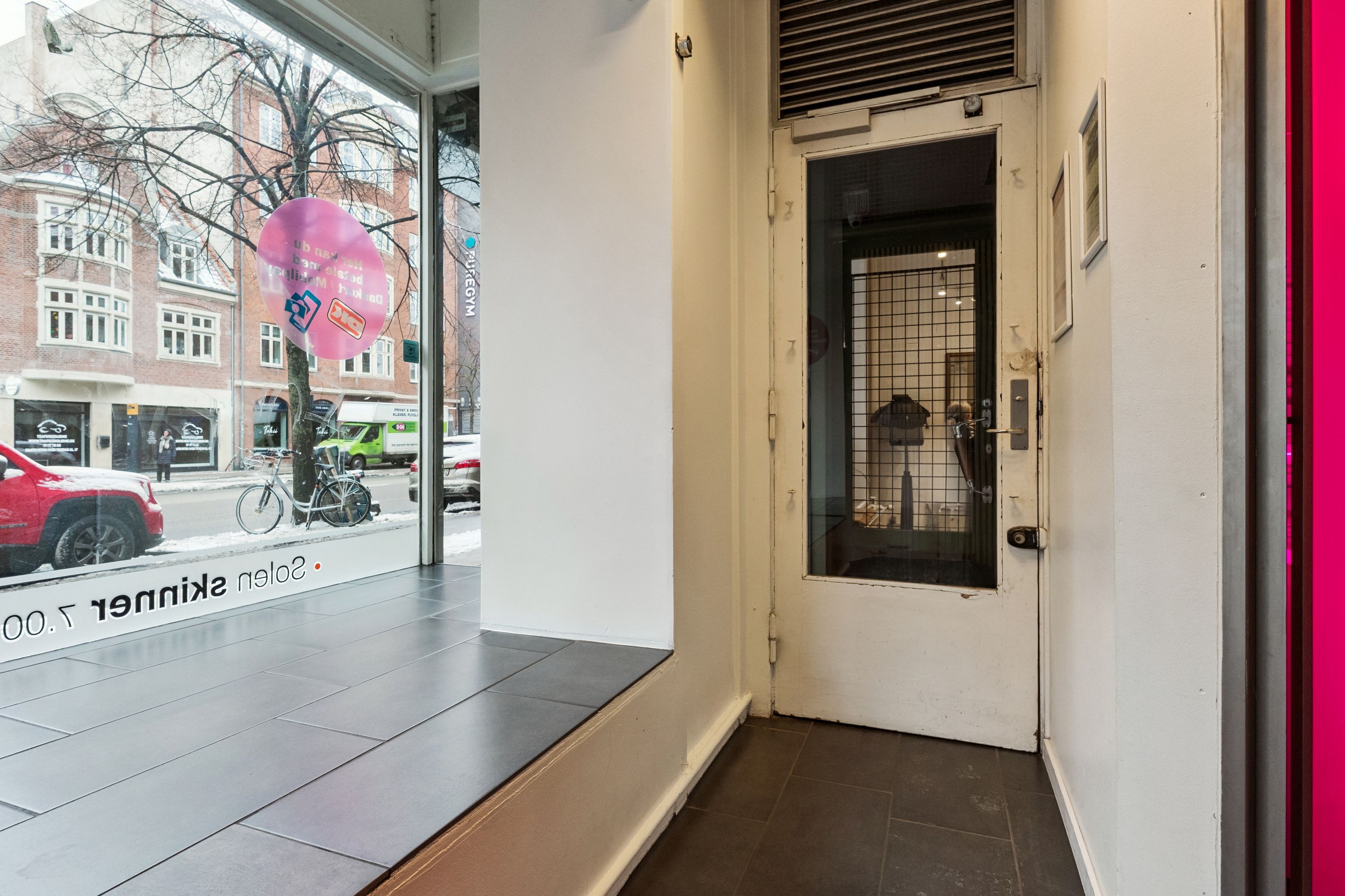 Butiksvindue med reklame og skrivning. En bil og cykel udenfor og en sølvfarvet dør med ruter. En række lyserøde og hvide vægge og en sølvfarvet dørhåndtag