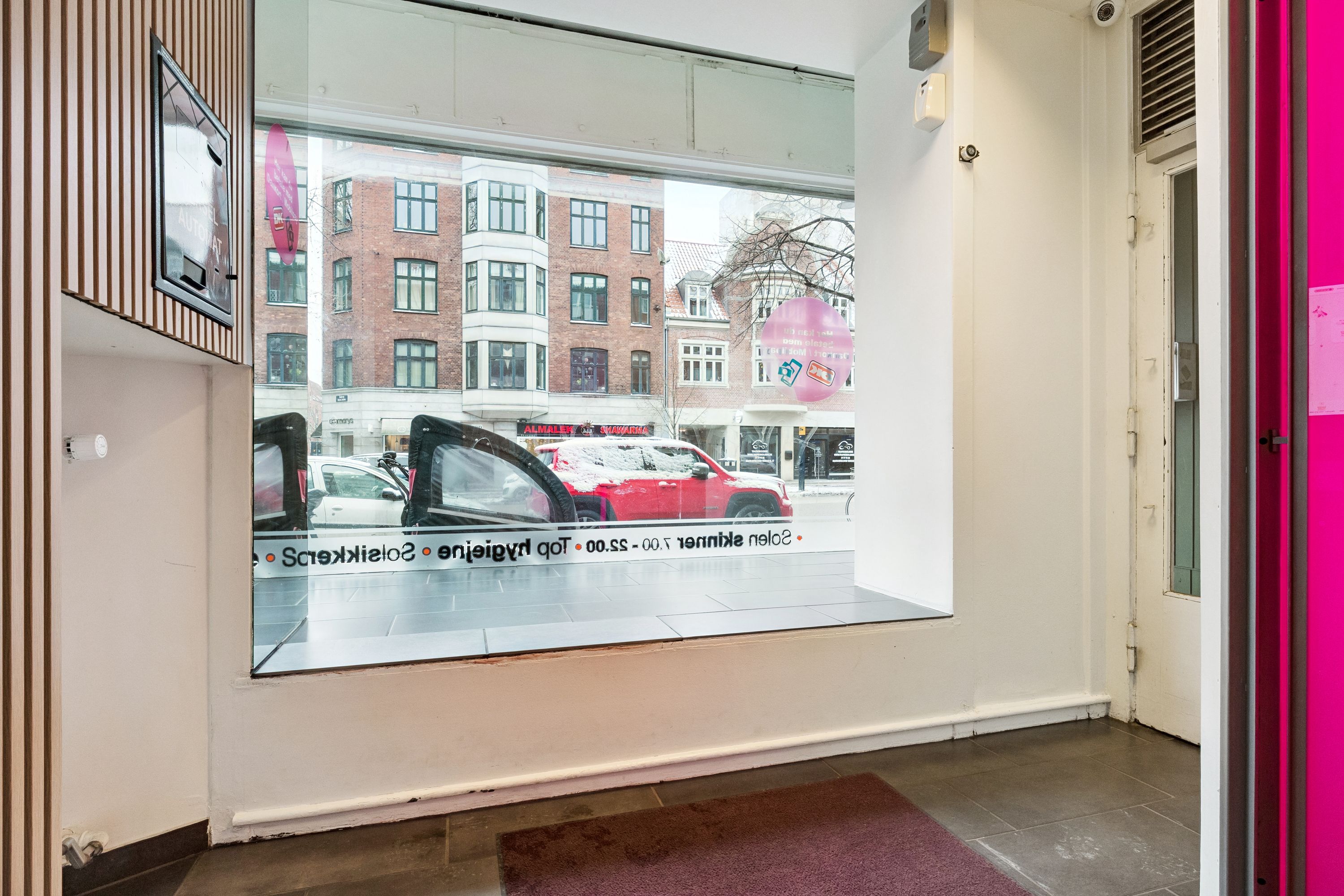 En butik til leje på Jagtvej 70 med vinduer, der reflekterer bygninger og biler, og indgangsdør med lyserosa tærskeler og indvendige vægge