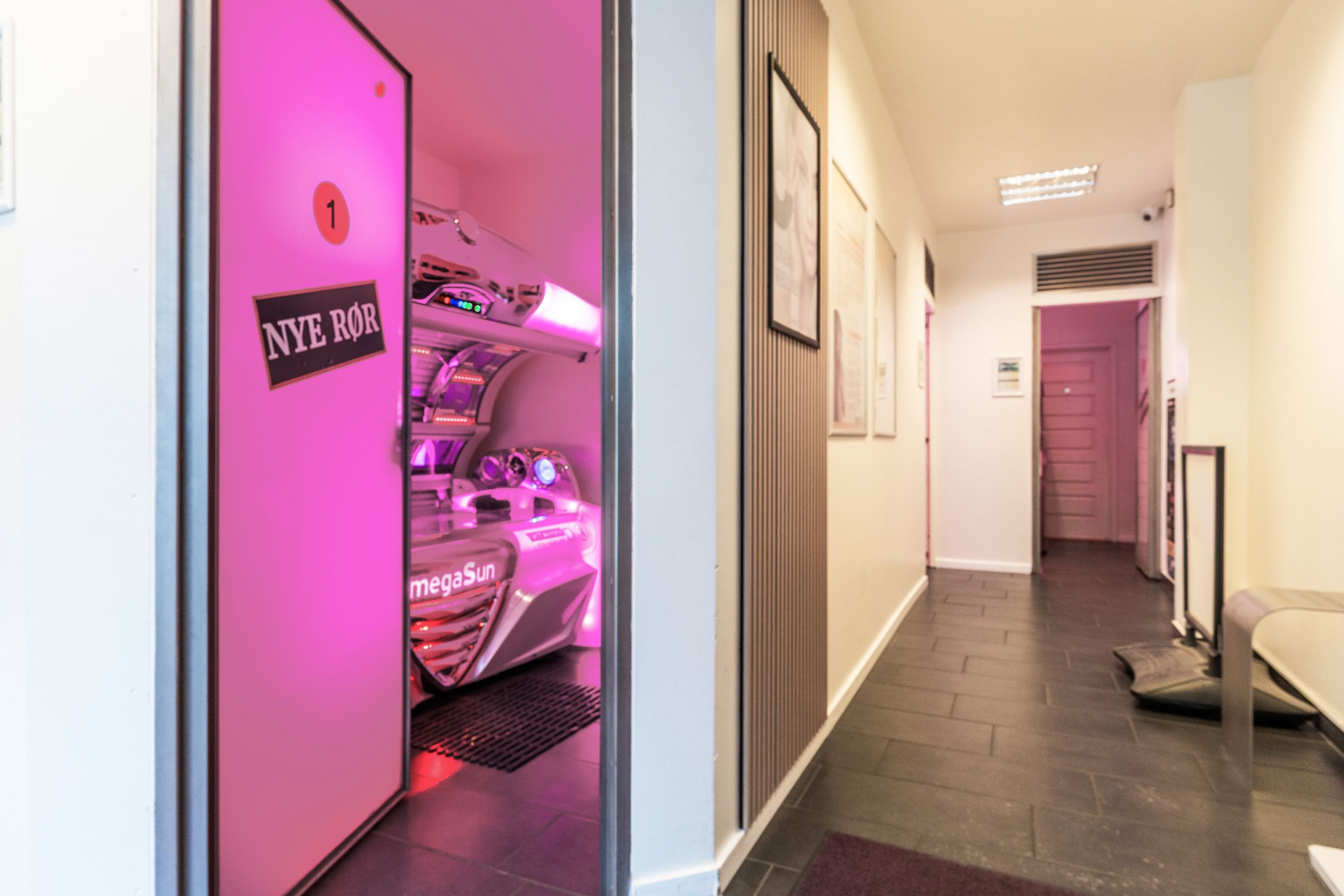 Pink og hvid solseng i butikken. Lyserødt vægpanel med billeder. Mørkt gulv med tegneserie på højre side. Sort indgangsdør med anslagstavle