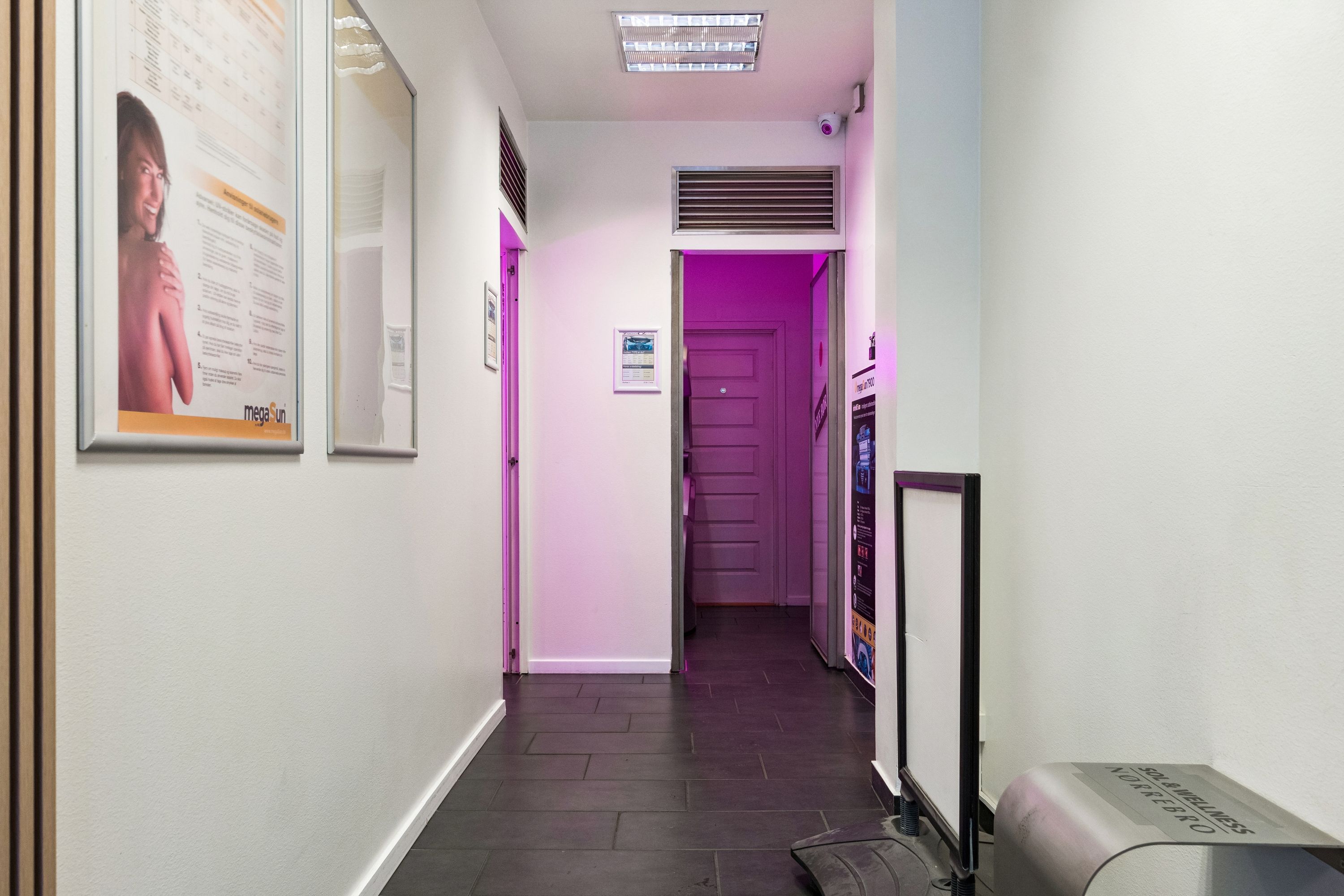 En indre gang i en moderne butik med hvide vægge og sort gulv, med en lys pink farve fra døren. Der er en reklameplakat med en kvinde og en tekst på Mega Sun.