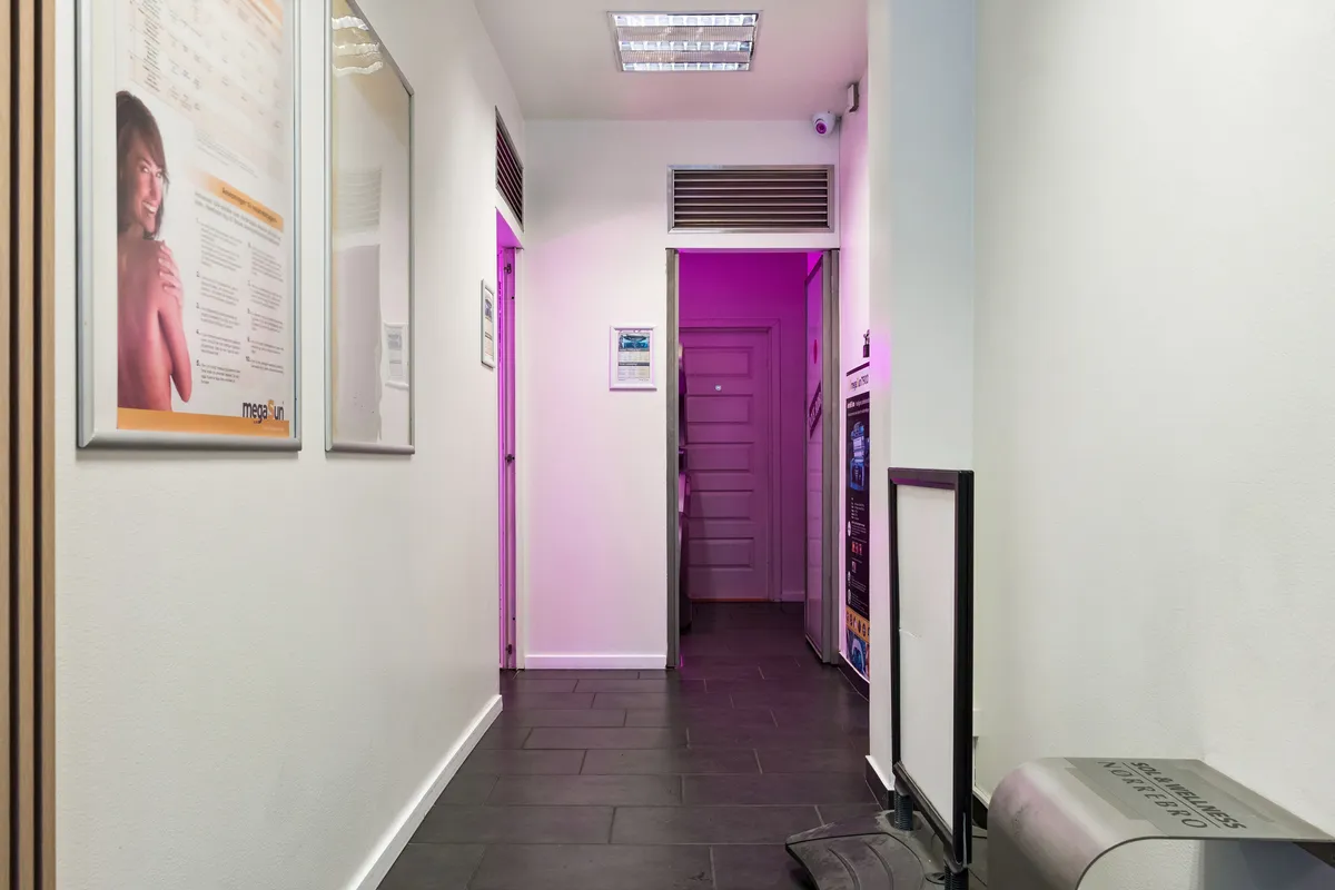 En indre gang i en moderne butik med hvide vægge og sort gulv, med en lys pink farve fra døren. Der er en reklameplakat med en kvinde og en tekst på Mega Sun.