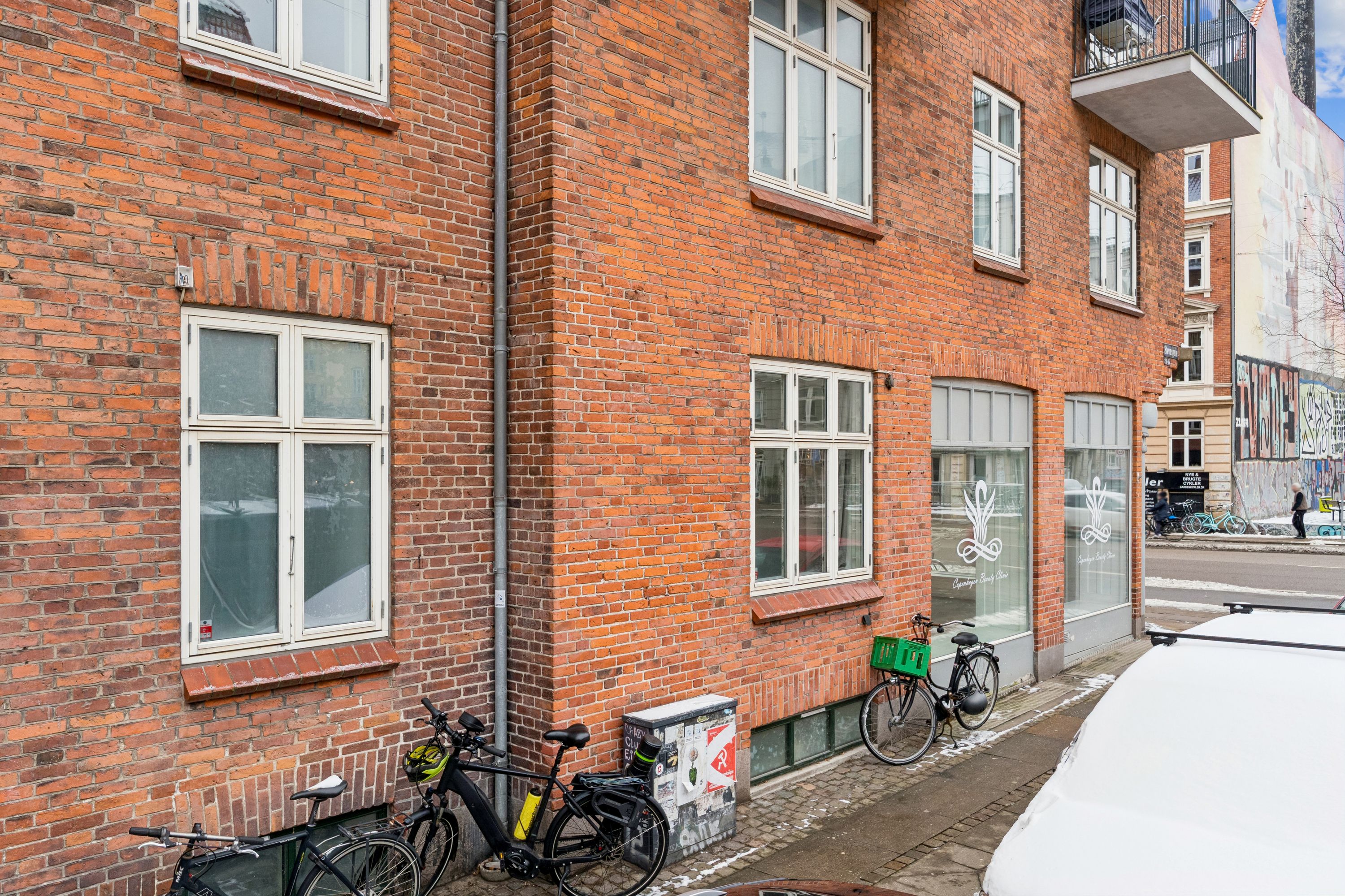 Rød murstensbygning med tre vinduer og to cykler parkeret udenfor. En anden cykel med kurv og en bil med sne foran. Butik med skilt og glasdør.