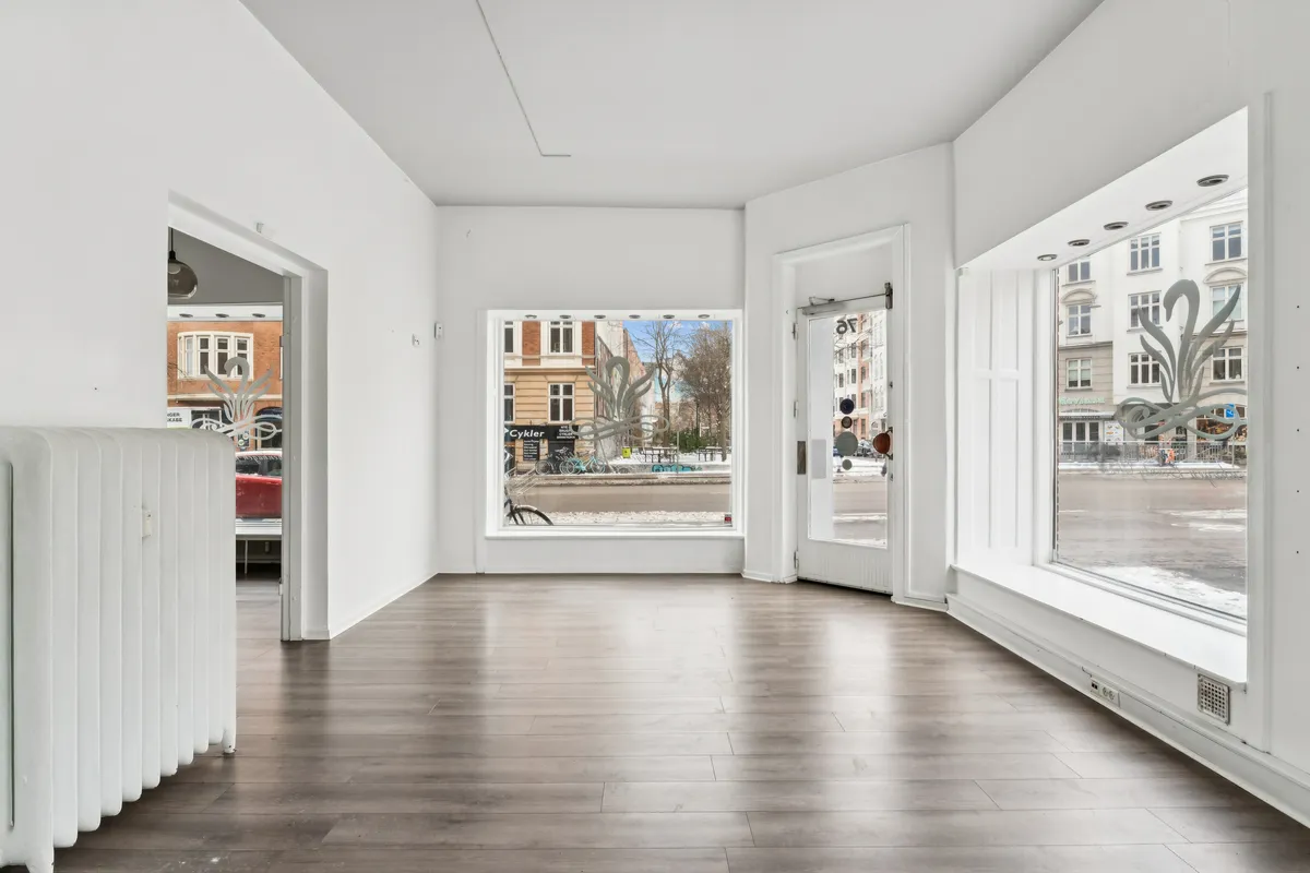 Store lyse vinduer med smukke mønstre, hvide vægge, elegant radiator og trægulv i en butik med udsigt til en bilparkering og gaden