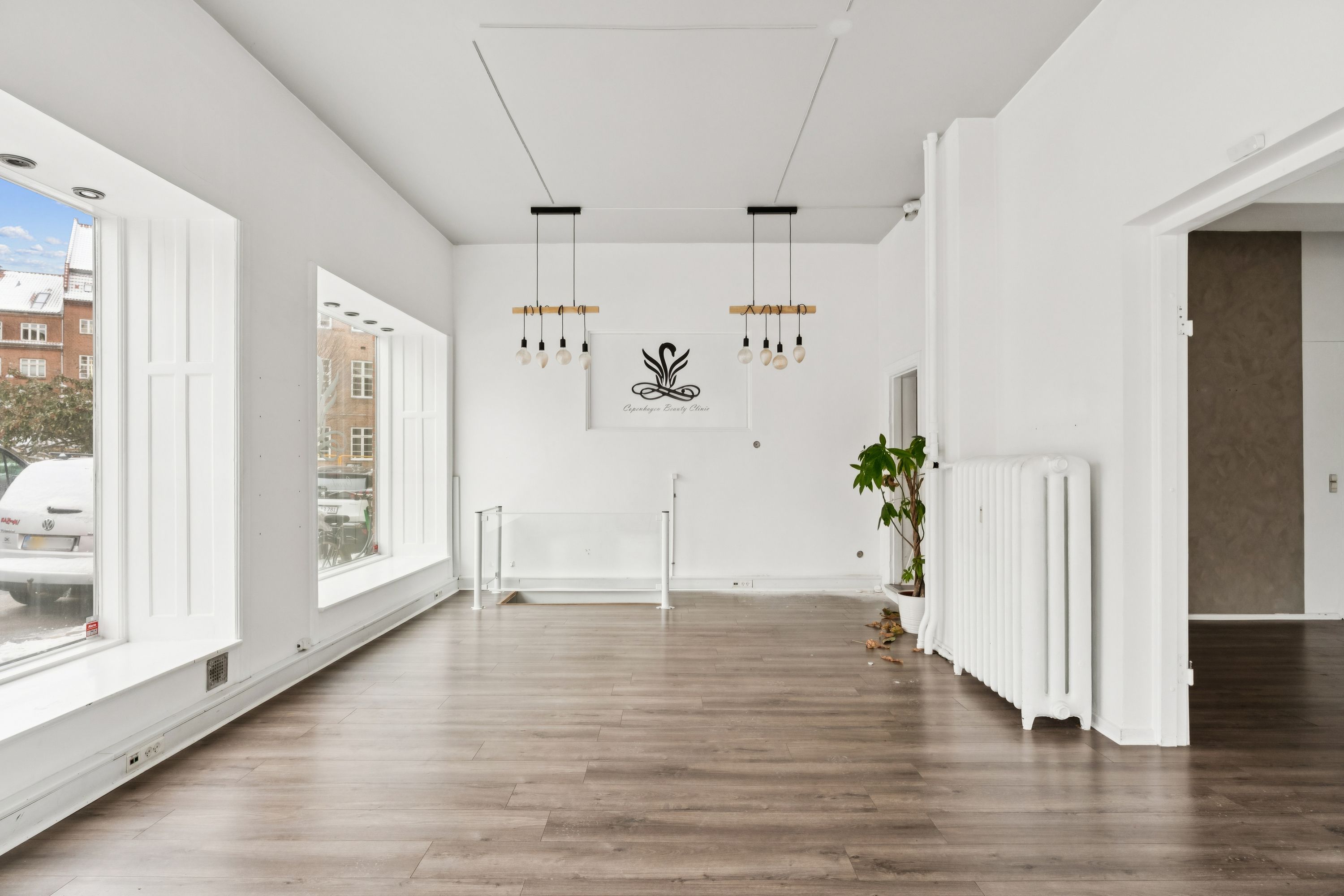 En stor og lys møbleret butikslokale med hvidt loft, gulv og vægge samt store vinduer og moderne lysekroner samt en plante og et hvidt radiator