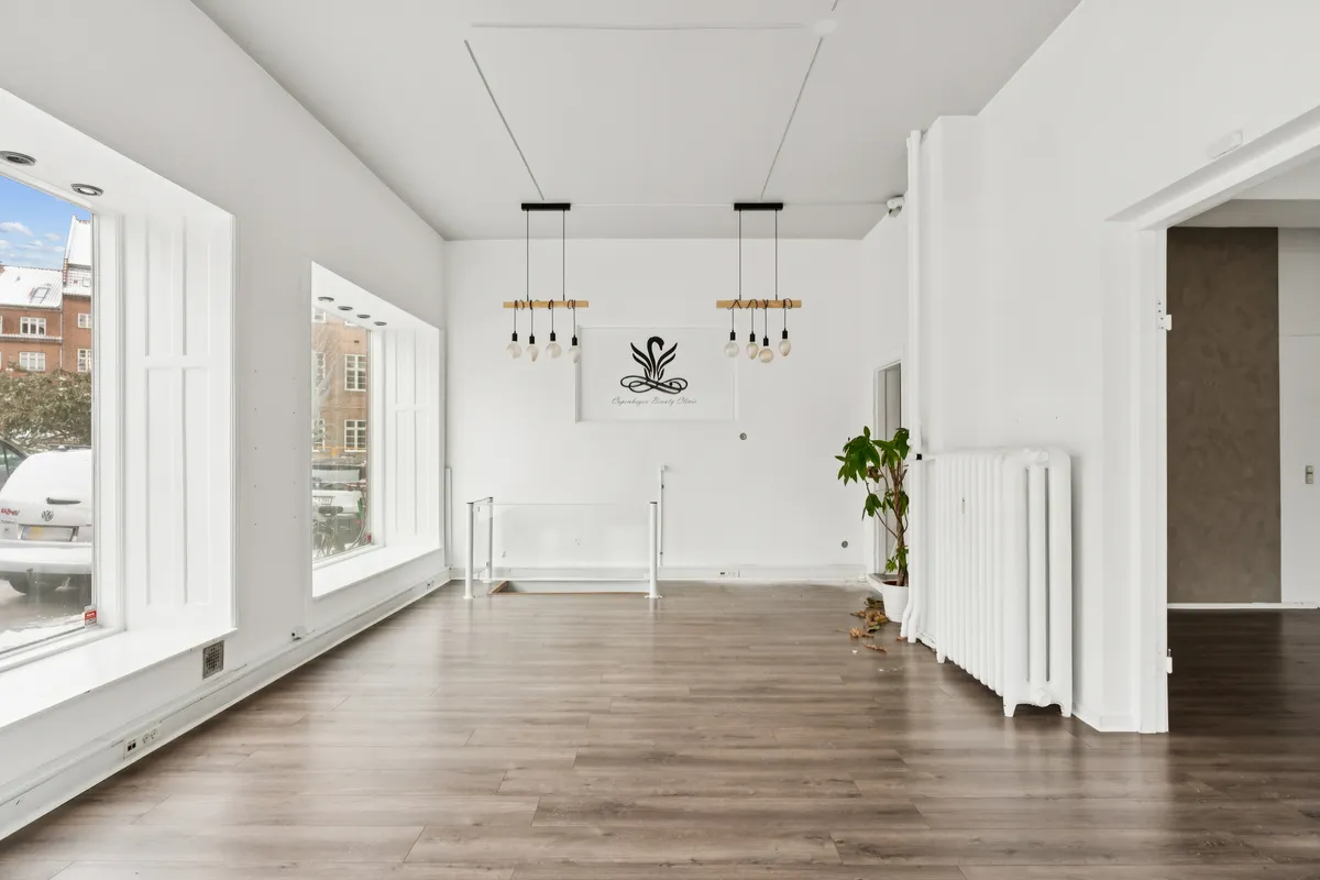 En stor og lys møbleret butikslokale med hvidt loft, gulv og vægge samt store vinduer og moderne lysekroner samt en plante og et hvidt radiator