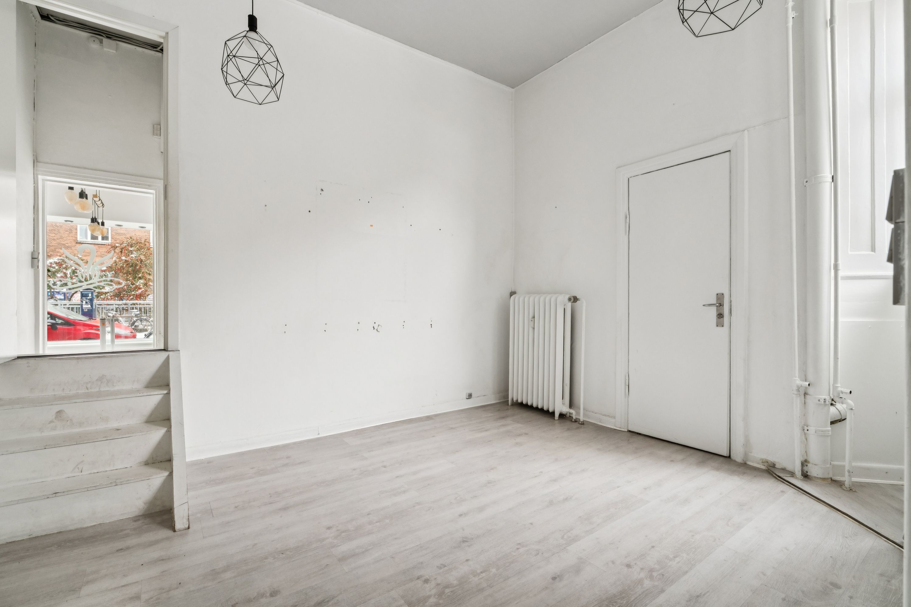 Lejlighed med hvidt loft, vægge og gulv, og en hvid radiator. Vindue med udsigt til en rød bil og bygninger samt en hvid dør med sølvfarvet håndtag