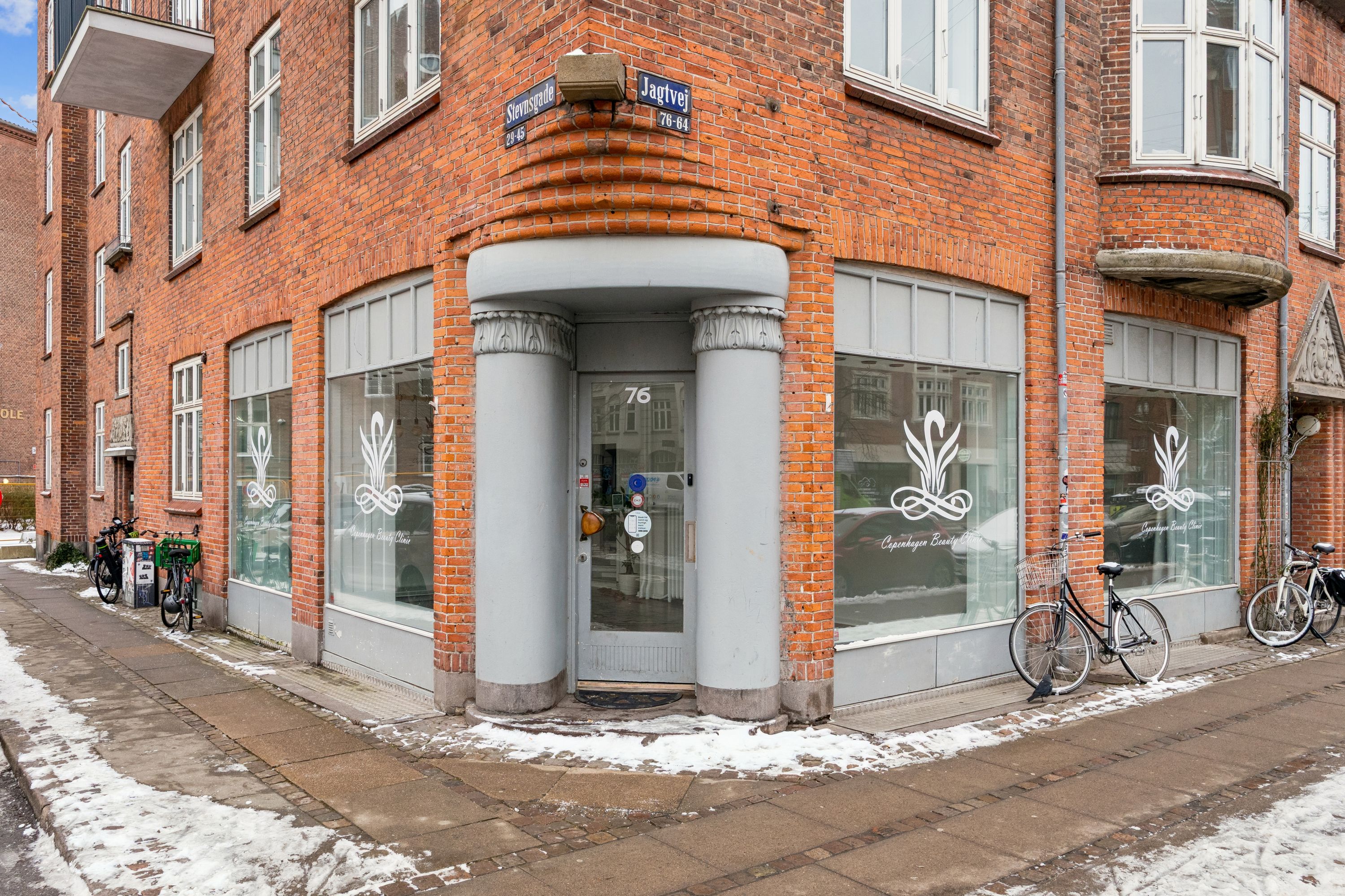 Butik i København med glasruder, logo, og nummer 76 på Jagtvej, med sne og cykler på fortovet og røde mursten.