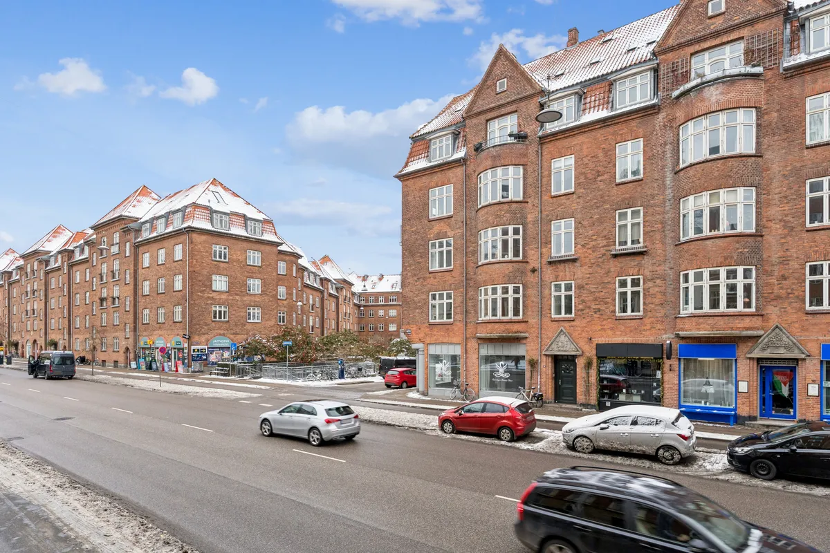 Butikstilbud på Jagtvej 76, 2200 København N. Tre biler kører på vejen, og tre biler er parkeret. Butikslokaler med glasvinduer. Sne på tømmer og biler.