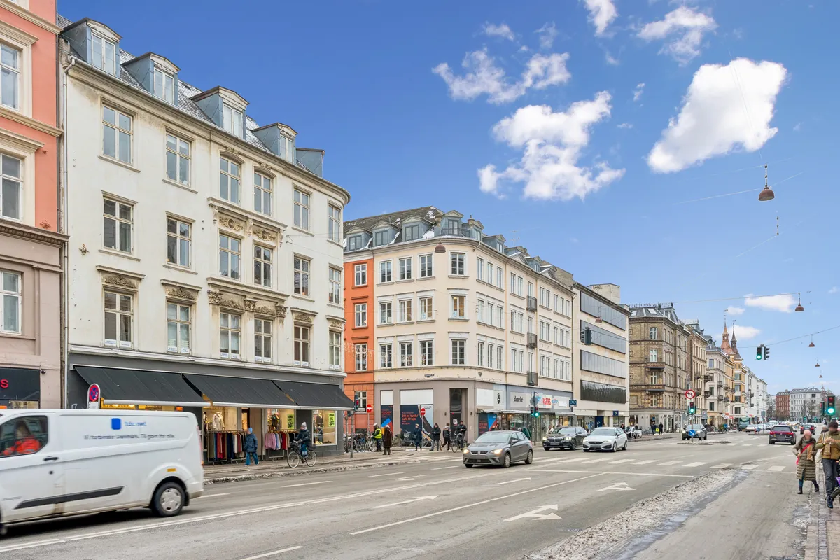 En bil kører gennem en bygade med mennesker og cyklister på forsiden og bygninger med butikker og gafler bagved