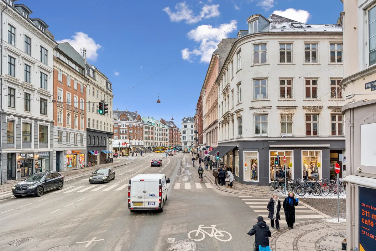 Mennesker går på gågaden, og der er biler og lastbiler på vejen. Der er trafiklys, bygninger og snesevis af cykler parkeret foran en bygning.