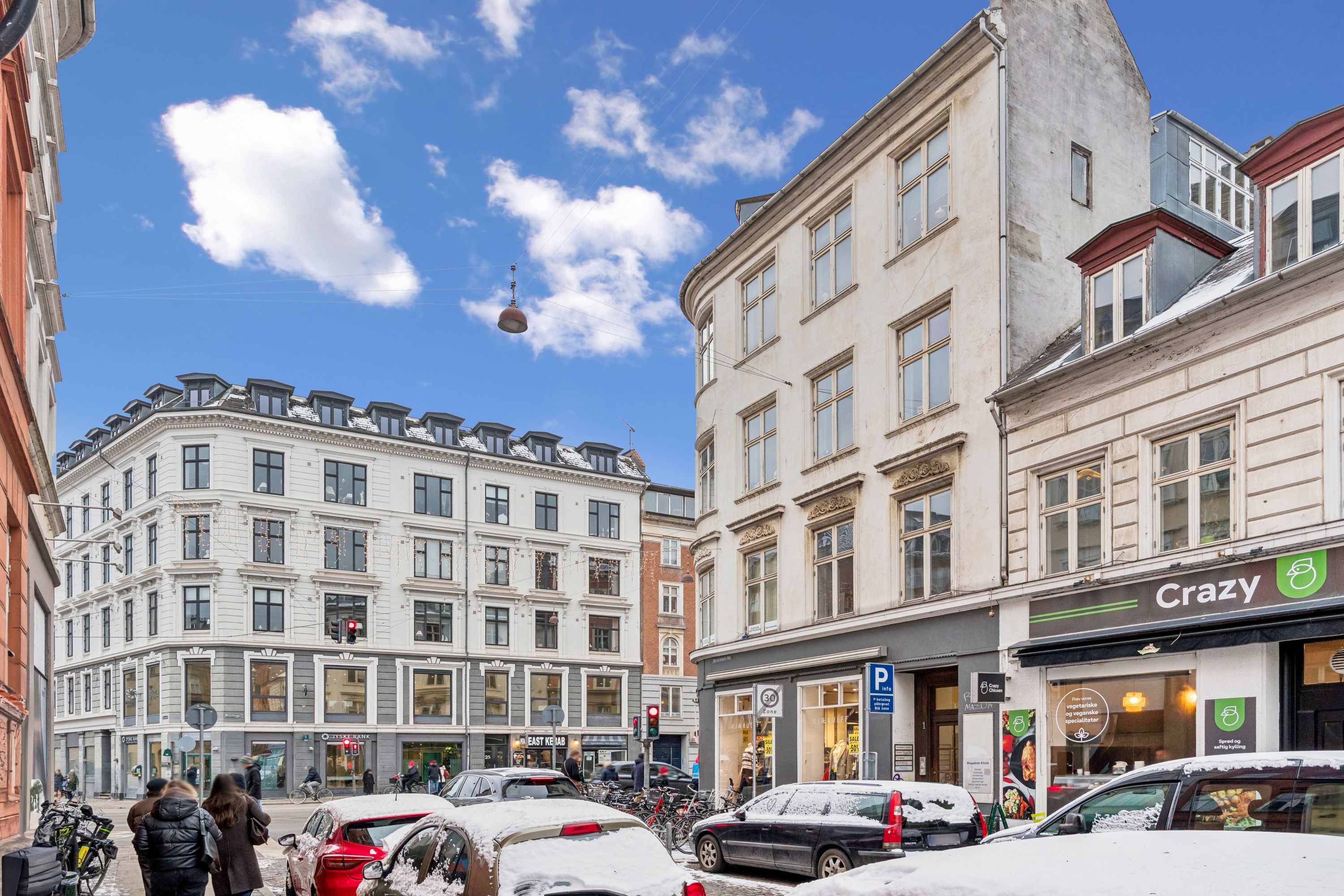 Strøget med biler og folk, der går forbi. Farverige bygninger og et skilt med ordet Crazy. Højt op hænger et skilt med ordet Bank.