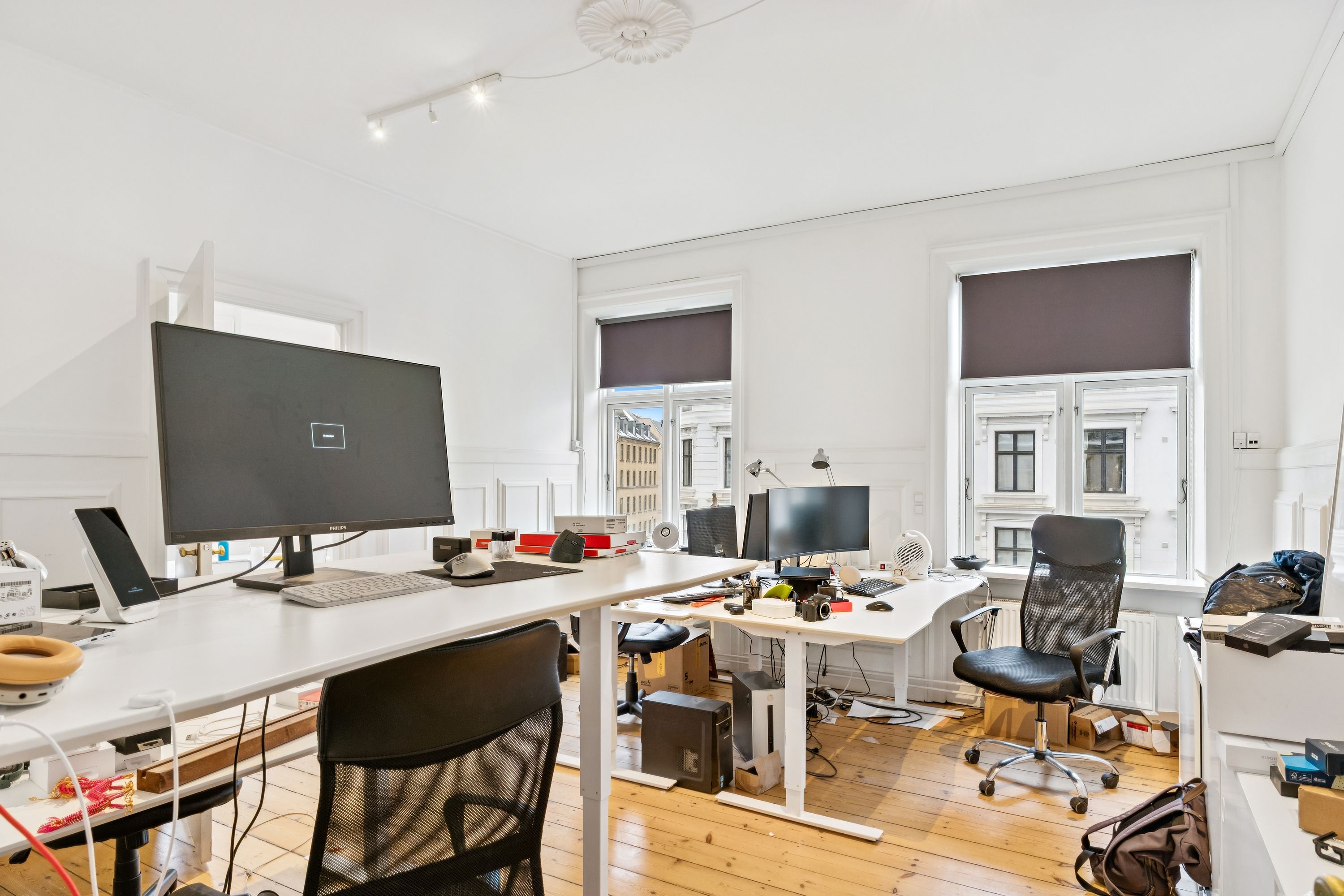 Kontor med to skriveborde, hver med en stående og en siddende computer, tæt på to store ruder med skyggegardiner og udsigt til en bygning med mange vinduer