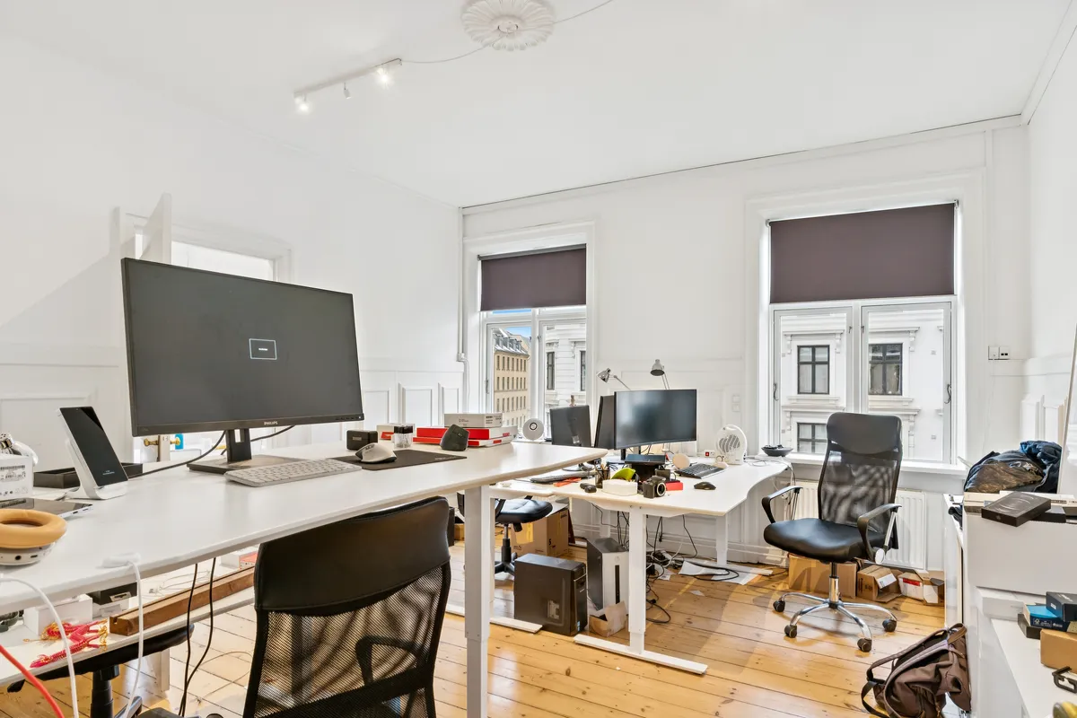 Kontor med to skriveborde, hver med en stående og en siddende computer, tæt på to store ruder med skyggegardiner og udsigt til en bygning med mange vinduer