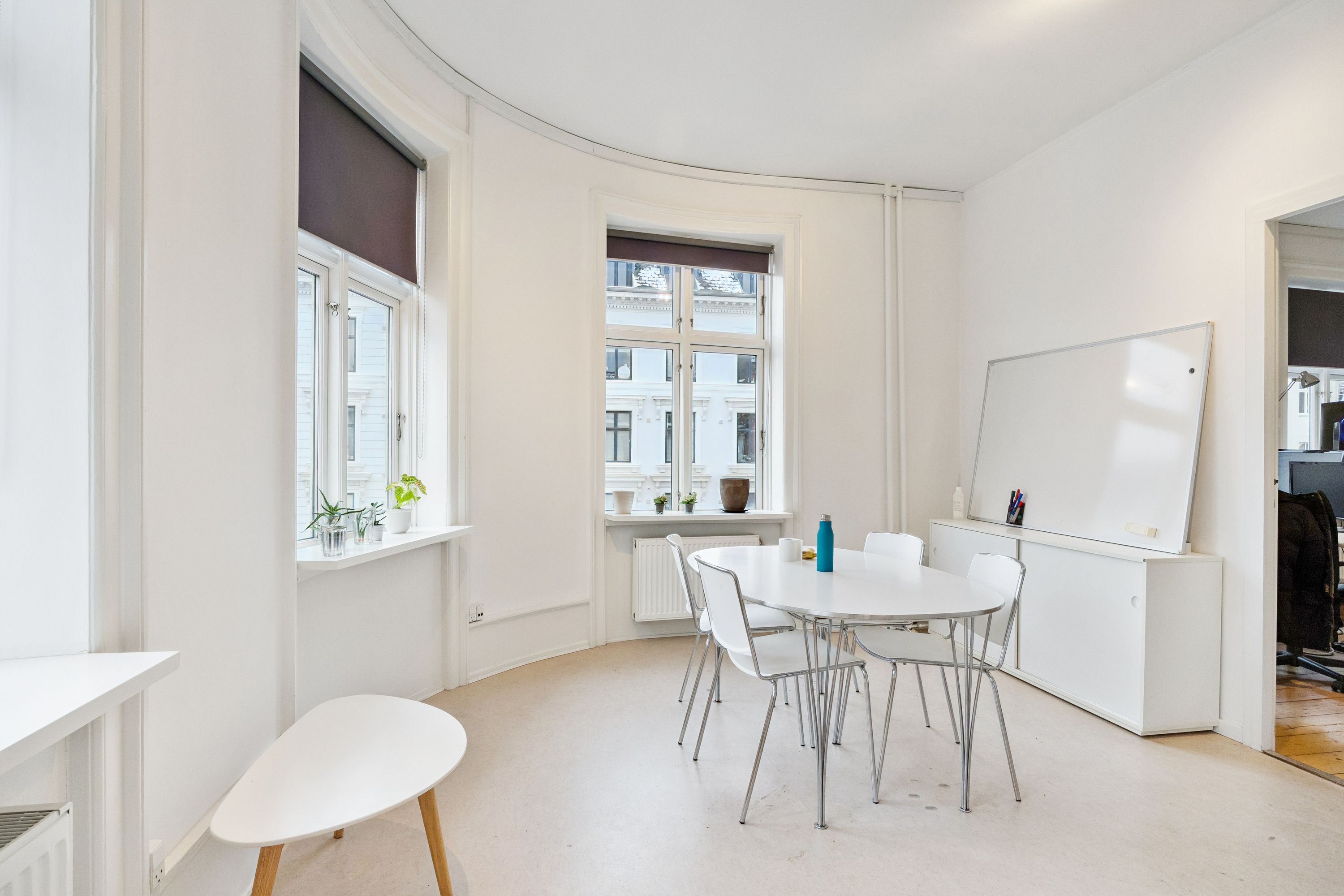 Lyst og moderne kontor med hvid væg, hvide stole og runde borde, lysevinduer med gardiner og små plantager, samt en hvid skrivebordsvæg med et hvidt tavle