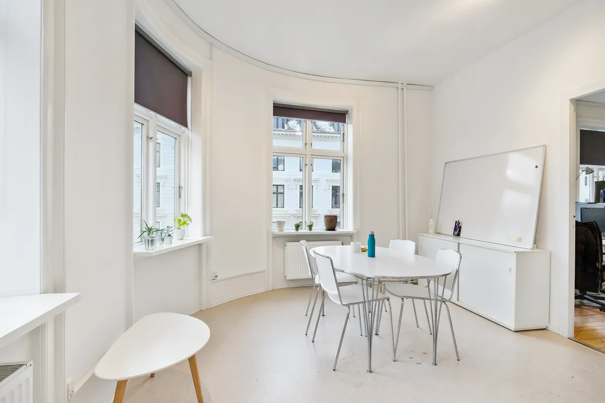 Lyst og moderne kontor med hvid væg, hvide stole og runde borde, lysevinduer med gardiner og små plantager, samt en hvid skrivebordsvæg med et hvidt tavle