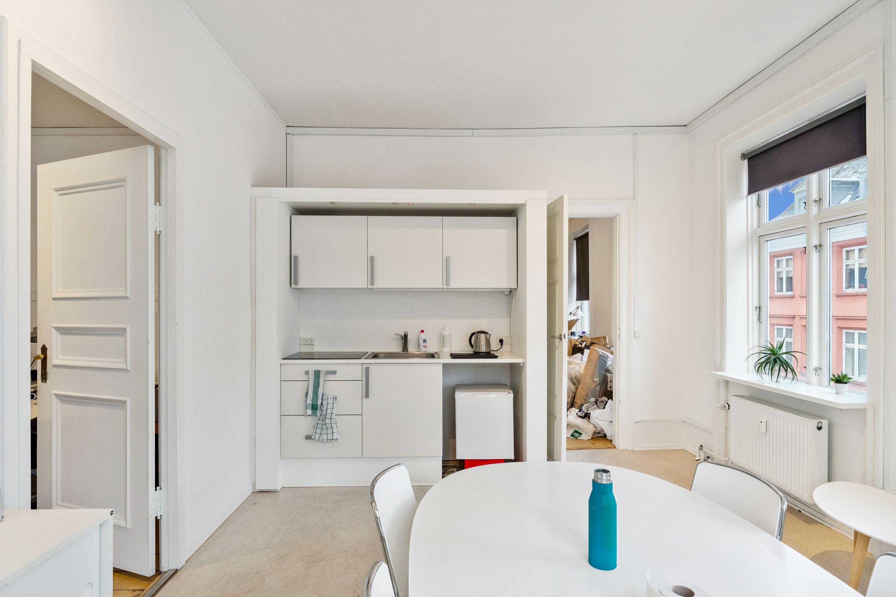 Moderne, lyse og minimalistisk indrettet køkken med hvide vægge, lysegrå gulv og runde spisebord med en blå vandflaske. Væggen har et stort vindue med gardiner og udendørs udsigt.