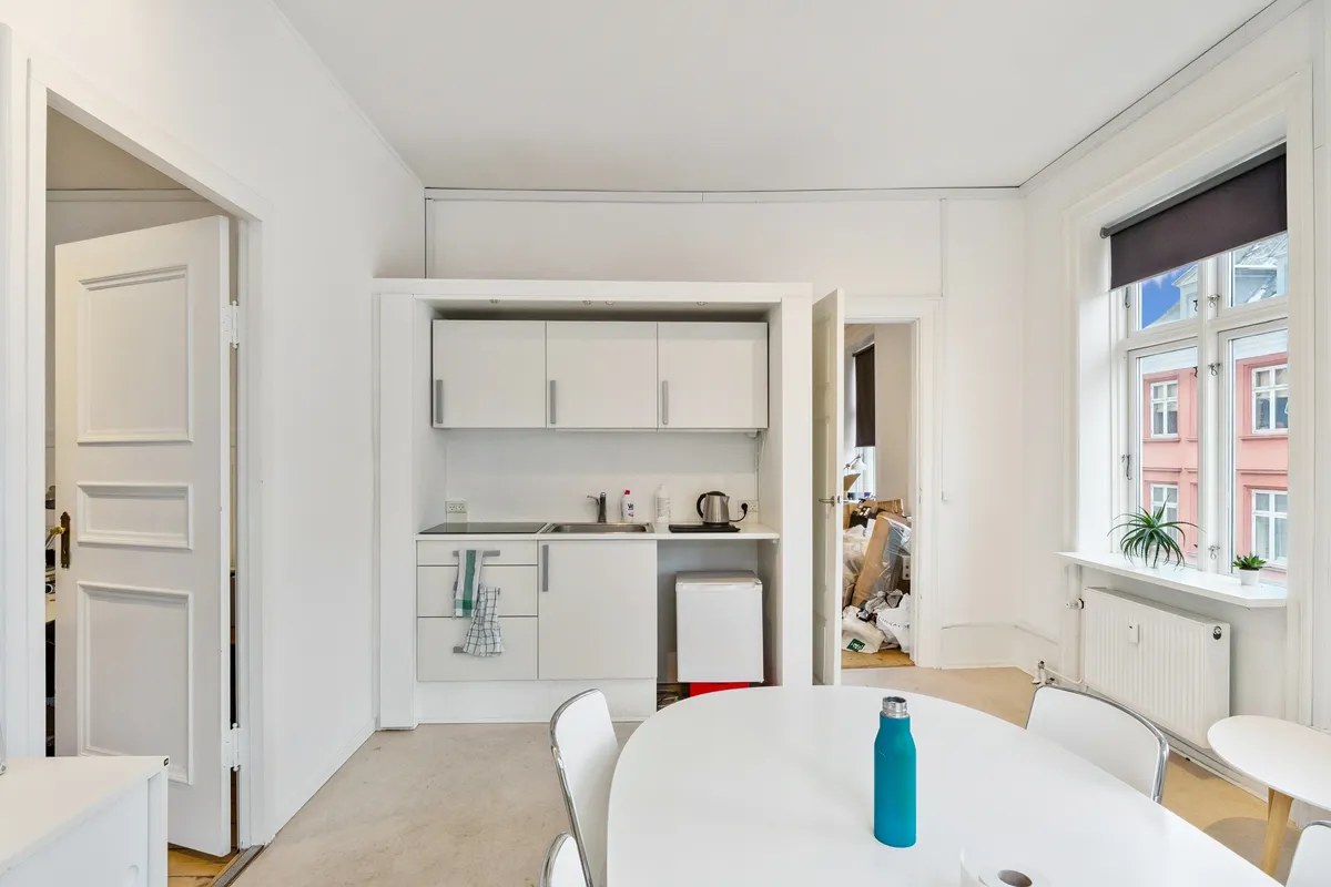 Moderne, lyse og minimalistisk indrettet køkken med hvide vægge, lysegrå gulv og runde spisebord med en blå vandflaske. Væggen har et stort vindue med gardiner og udendørs udsigt.