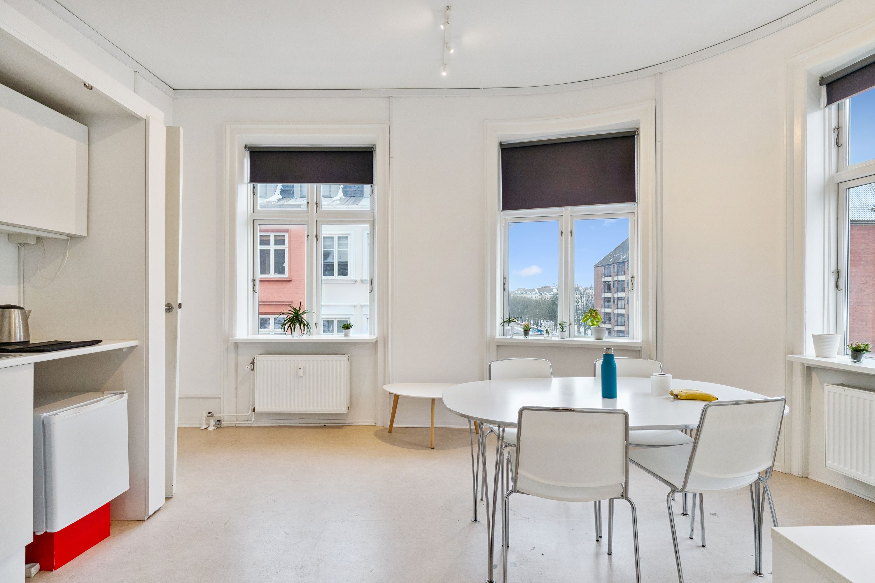 Kontor med lyse gulve og hvide vægge. Rundt bord med fire stole og en stol ved væggen. En flaske og en banan på bordet. Vindue med solskærme og udsigt til gaden med bygninger og planter