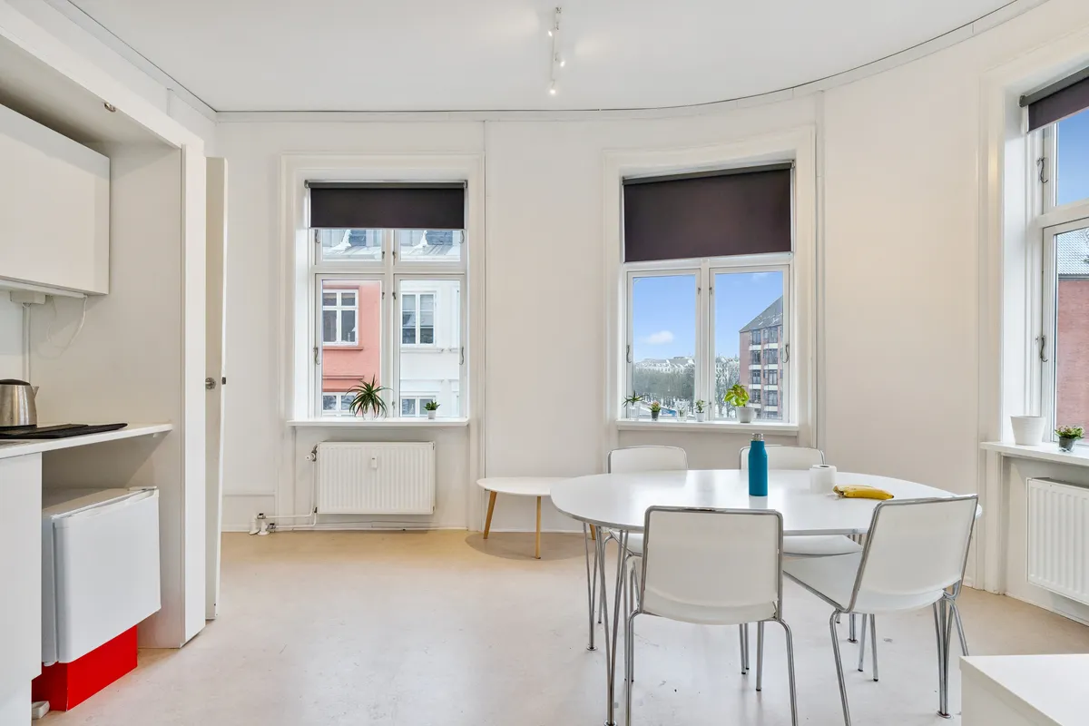 Kontor med lyse gulve og hvide vægge. Rundt bord med fire stole og en stol ved væggen. En flaske og en banan på bordet. Vindue med solskærme og udsigt til gaden med bygninger og planter