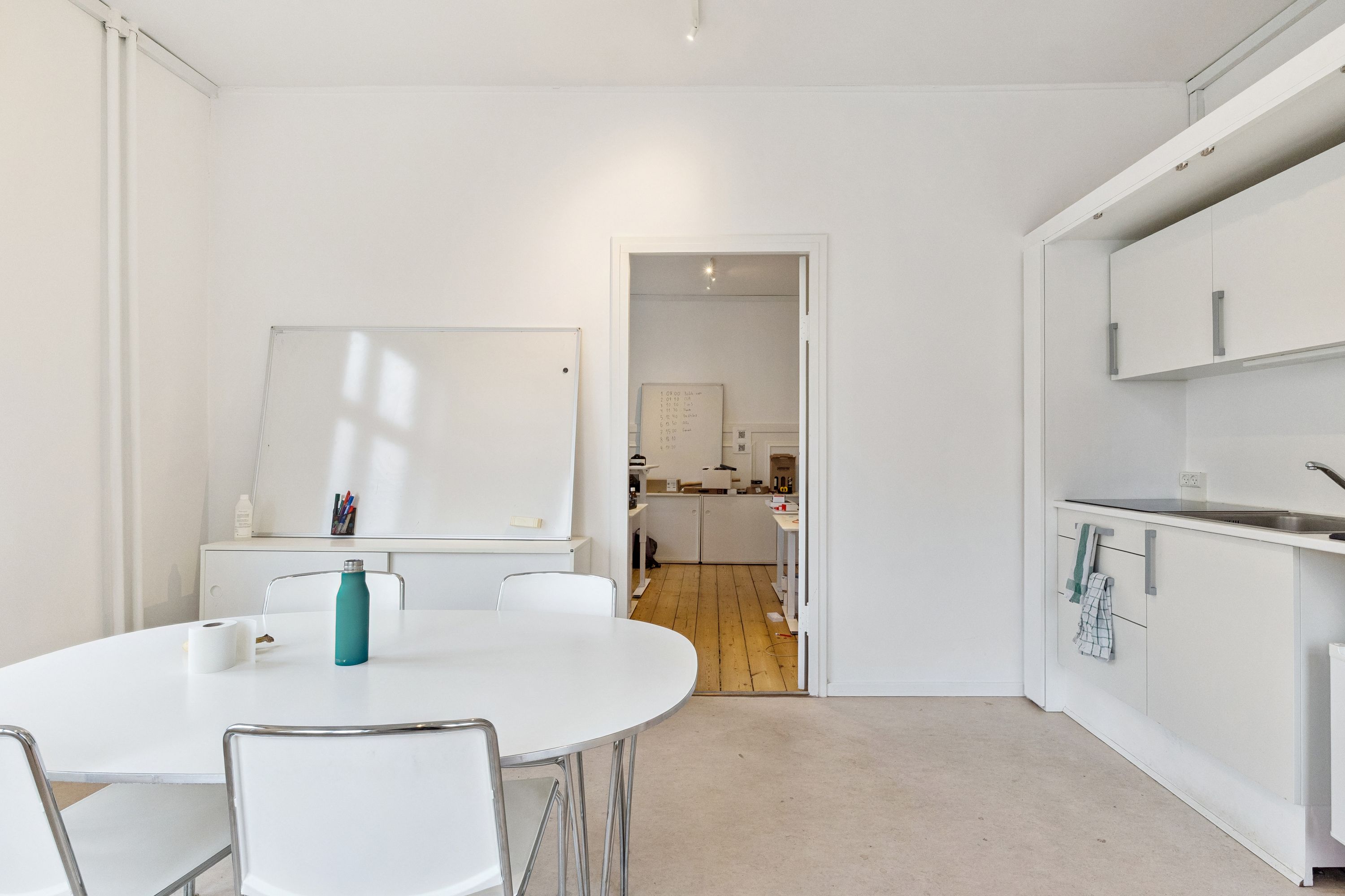 Kontor med hvide vægge, rundt bord med fire stole, lyse pladeborde, hvid tavle, en flaske, to ruller toiletpapir og en åben dør til køkken med hvide skabsfronter