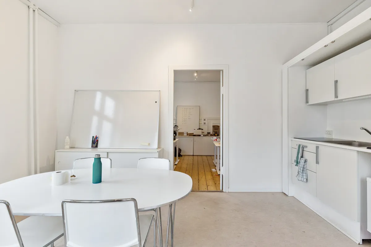 Kontor med hvide vægge, rundt bord med fire stole, lyse pladeborde, hvid tavle, en flaske, to ruller toiletpapir og en åben dør til køkken med hvide skabsfronter