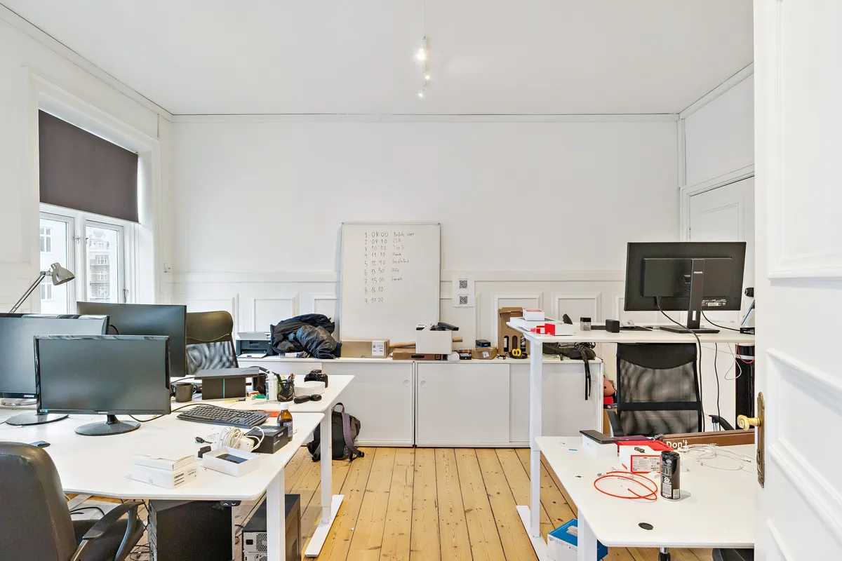 Kontor med to skriveborde, computere, skærme og skrivebordslamper samt en stol og skraldpose