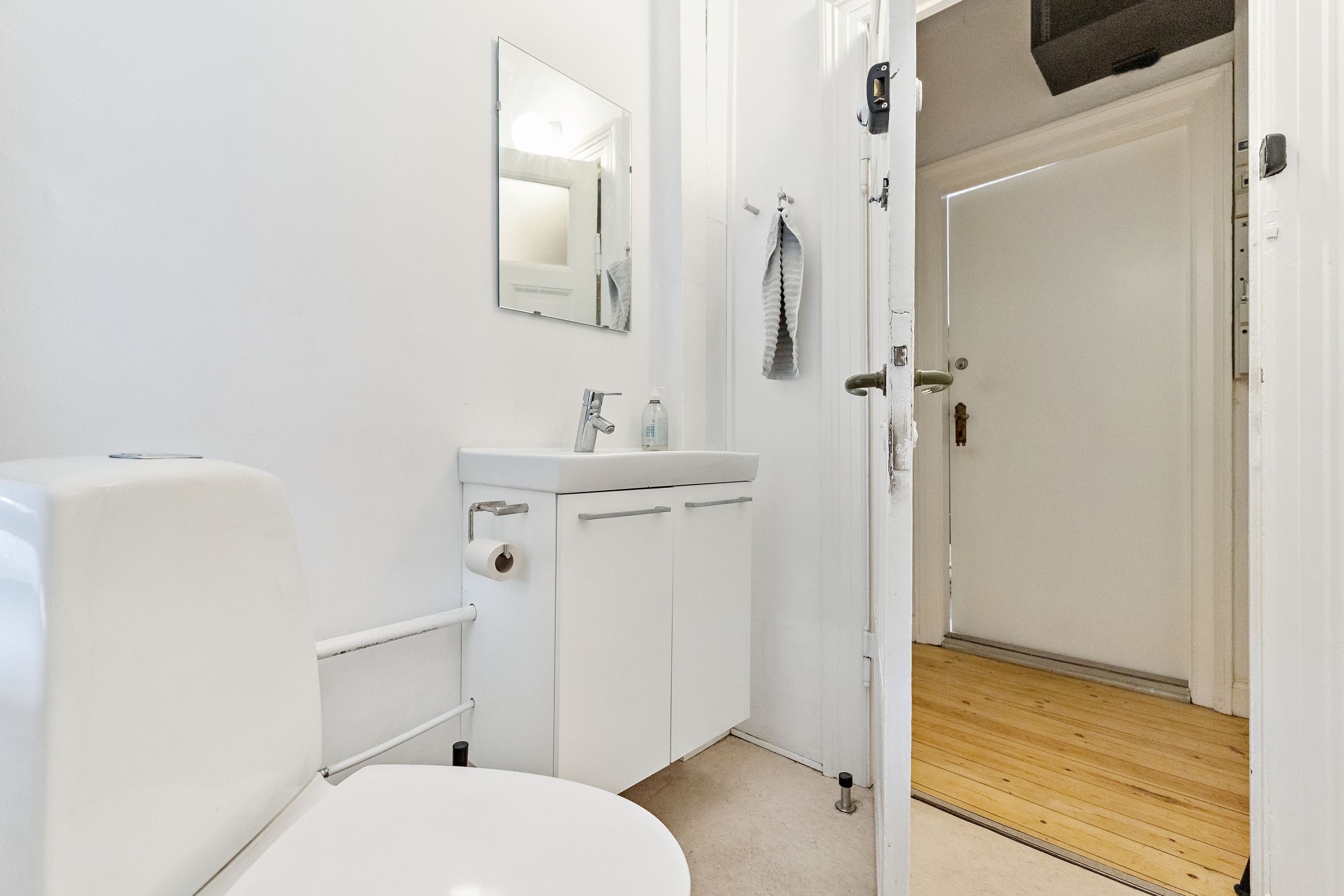 En moderne og pænt indrettet toilet med hvidt toilet, hvidt badeværelse med håndvask og spejl samt håndklæde i toiletrummet og trægulv i gangen til siden