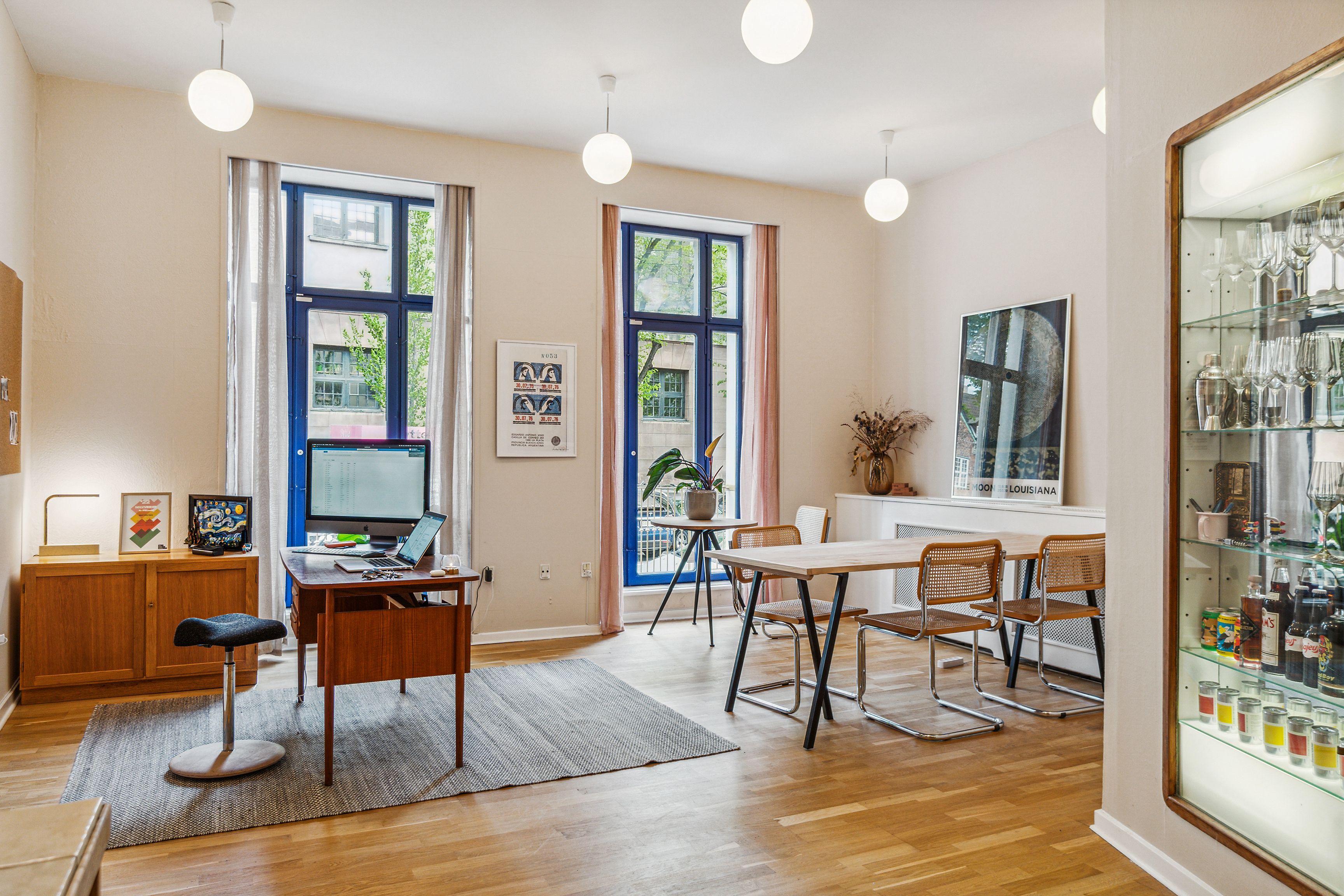 Kontor med skrivebord, computer, lampe og stole. Væg med hylde og maleri. Vindue med udsigt til byggede bygninger. Flad loft med hængende lampe.