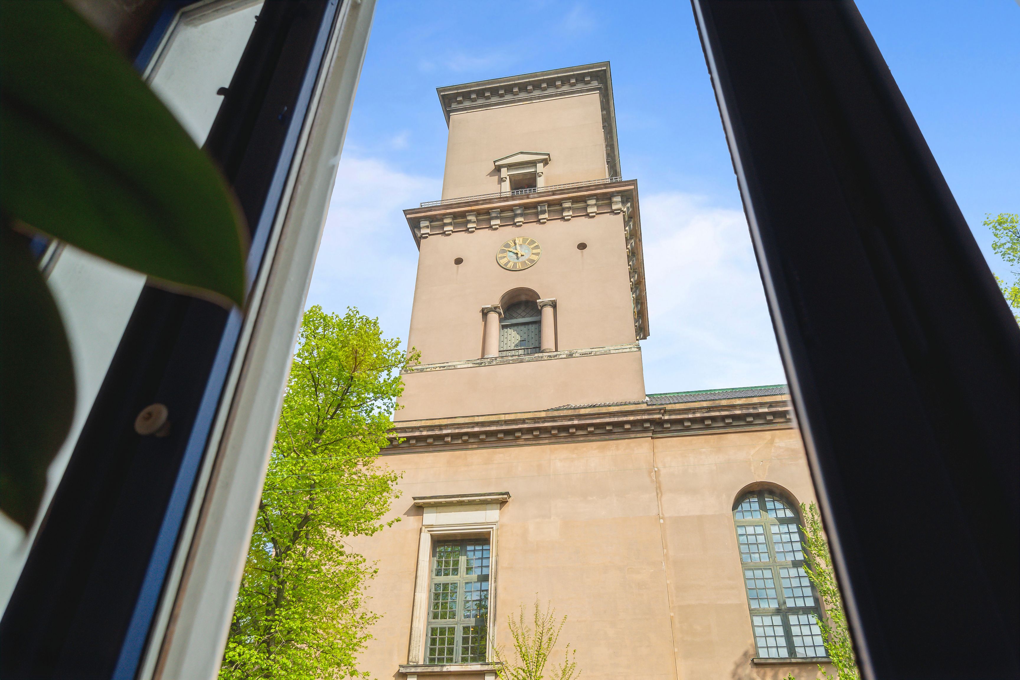 Kontor til leje på Dyrkøb 7 i København. Bygningen med klokketårn og lyse vinduer. Solskin og grønne træer med friske blade. Moderne og centralt beliggende.