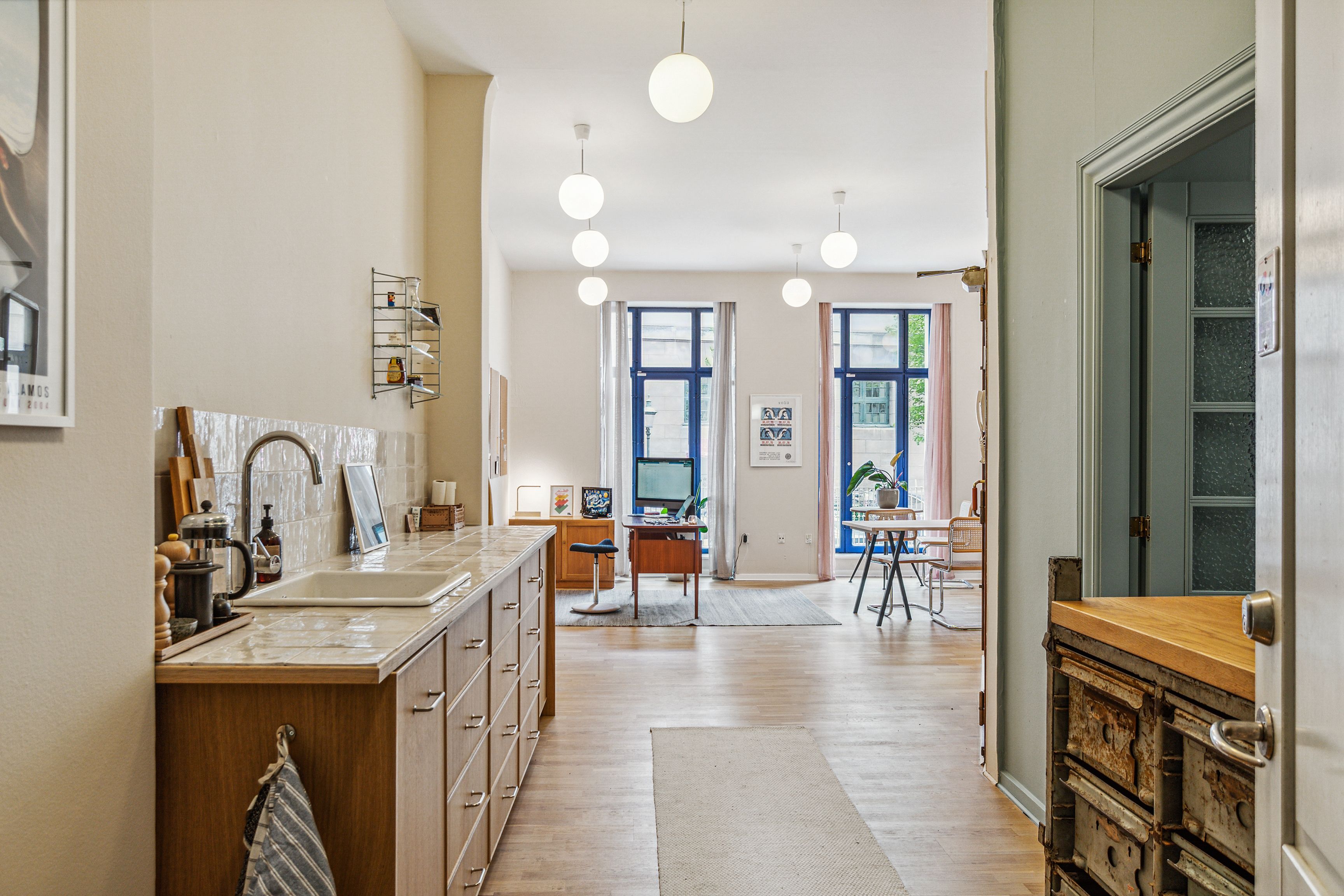 Kontor med køkken og siddekrog, moderne interiør, tre runde lysekroner, hvid keramisk gulv og træskabe med håndtag og flere skuffer.