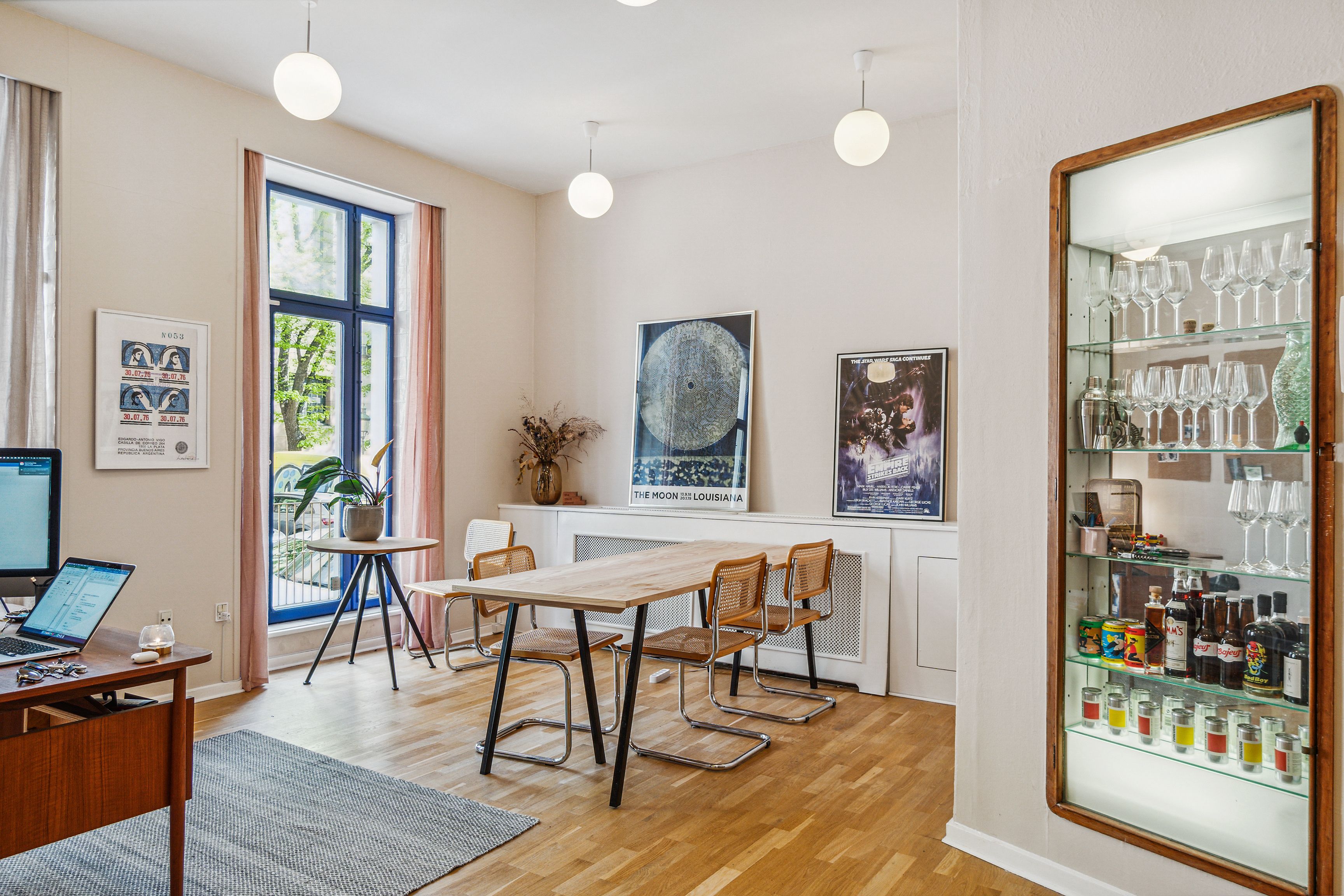 Kontor med højt til loftet, moderne møbler og lyse pærelampetter. En plante i krukken og et foto på væggen. En computer på bordet og en stol med høj ryglæn.