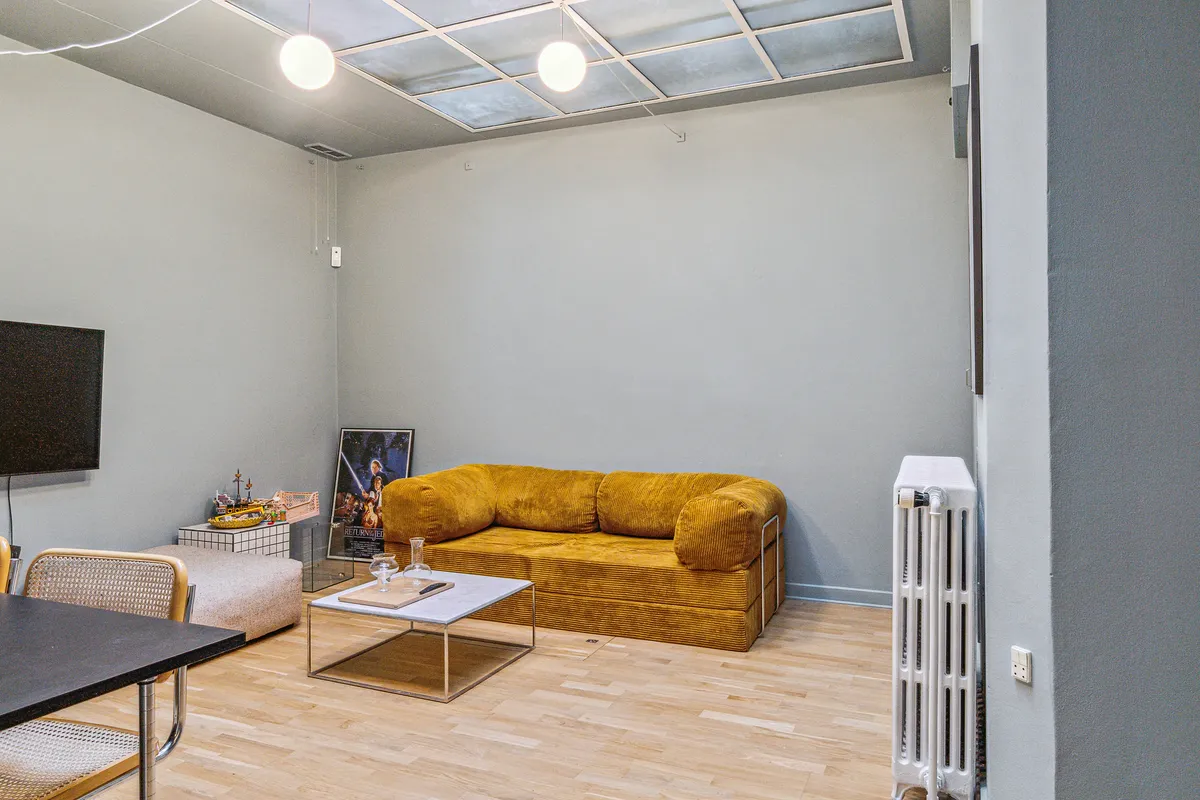 Kontor med møbler, sofa, kaffebord, tv, radiator og en Star Wars-plakat. Lys pære hængende fra loftet og lyseplader på loftet.