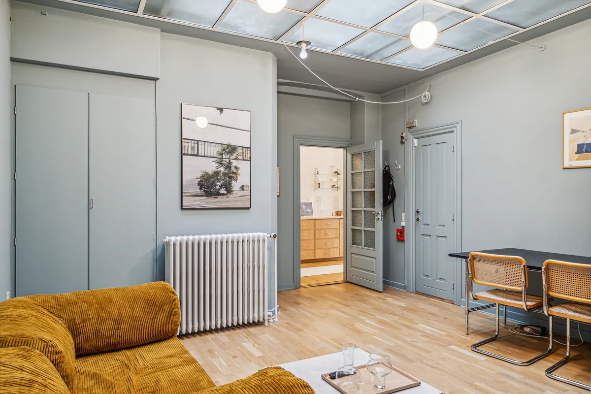Kontor med lyse lofter og radiator i en lejlighed. Møbler inkluderer en gul sofa og to lyse stole. Der er to malerier på væggen, en åben dør og en åben dør med et kammer.