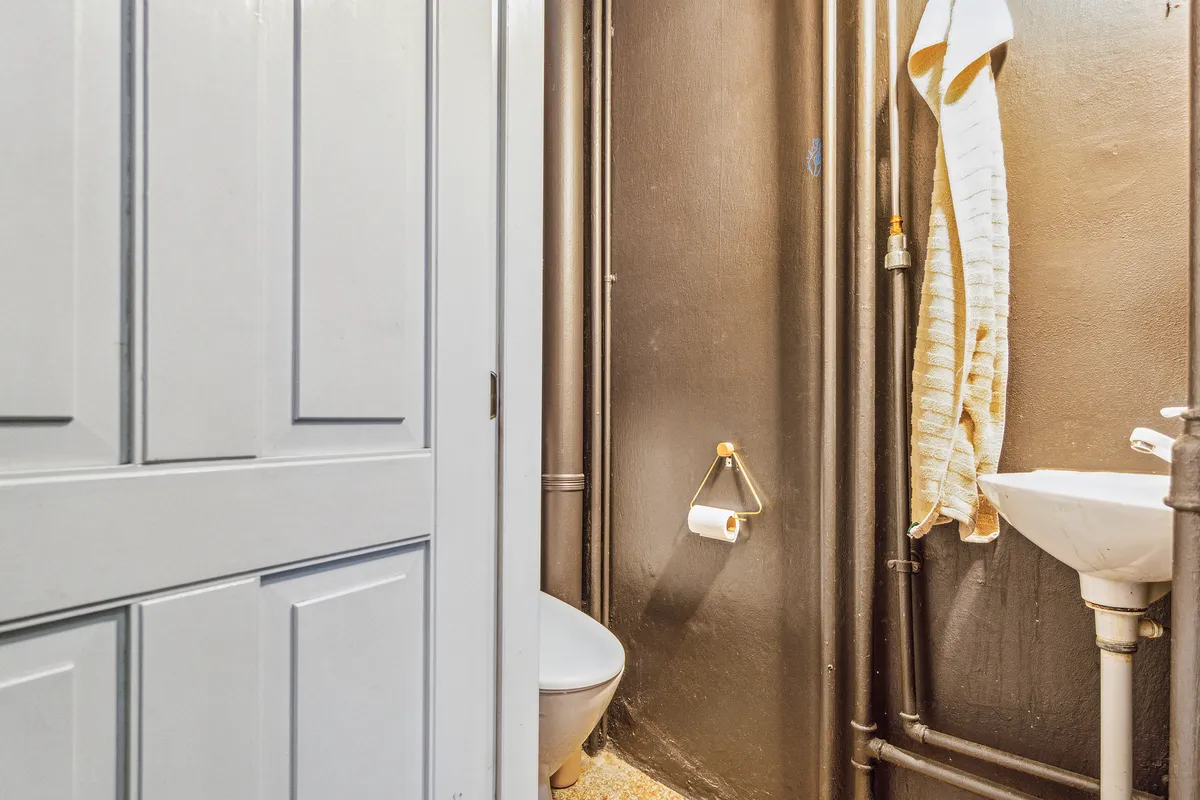 En lille badeværelse med hvidt toilet, rustfri metalrør og en hængende håndklæde. Toiletpapir er på toiletpapirholderen.