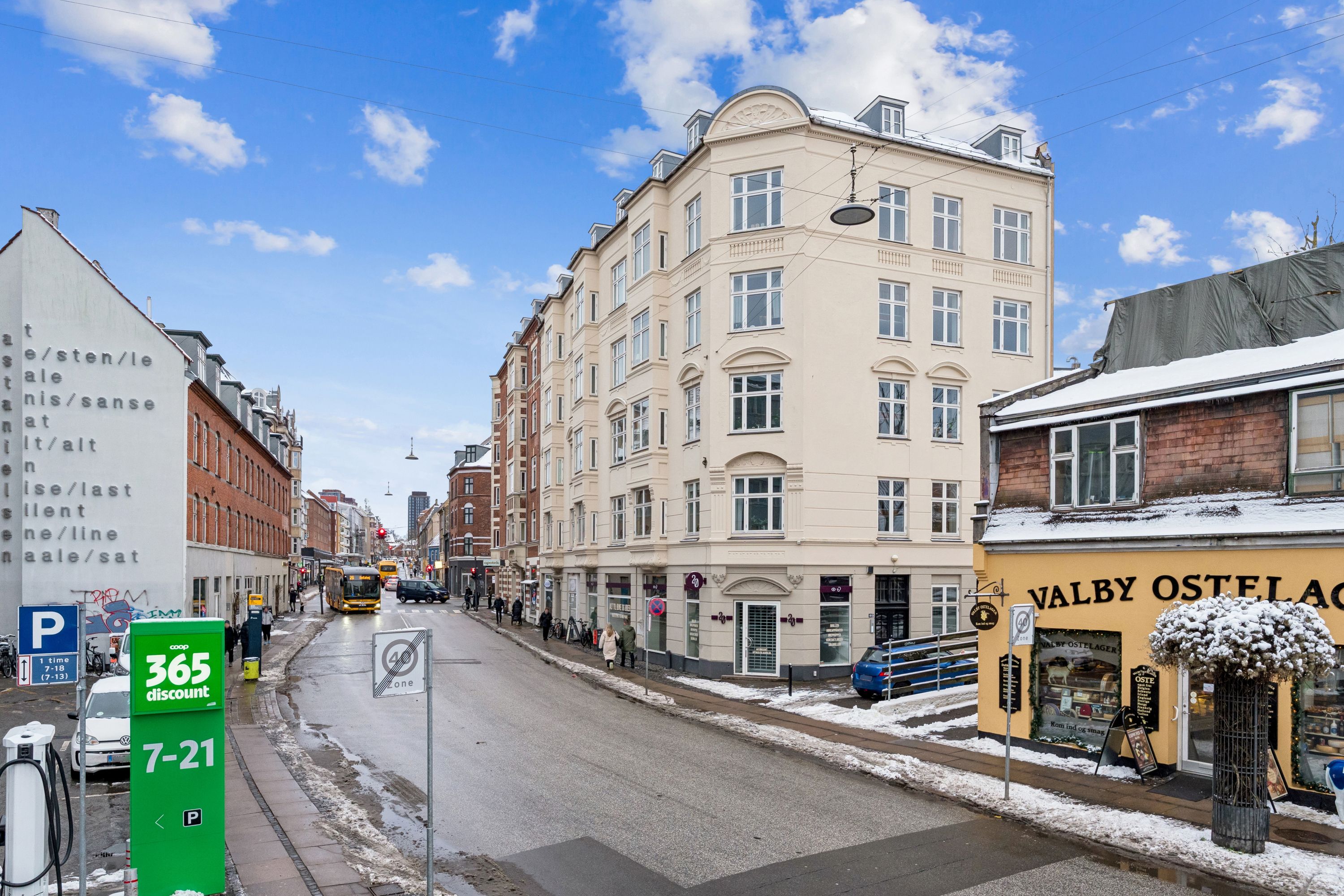 Markant hjørnebutik ved Valby Langgade 49B, st., med sne på taget og mennesker, der vandrer langs gaden, udstyret med elektriske parkeringsstationer og 365 discount-skilt