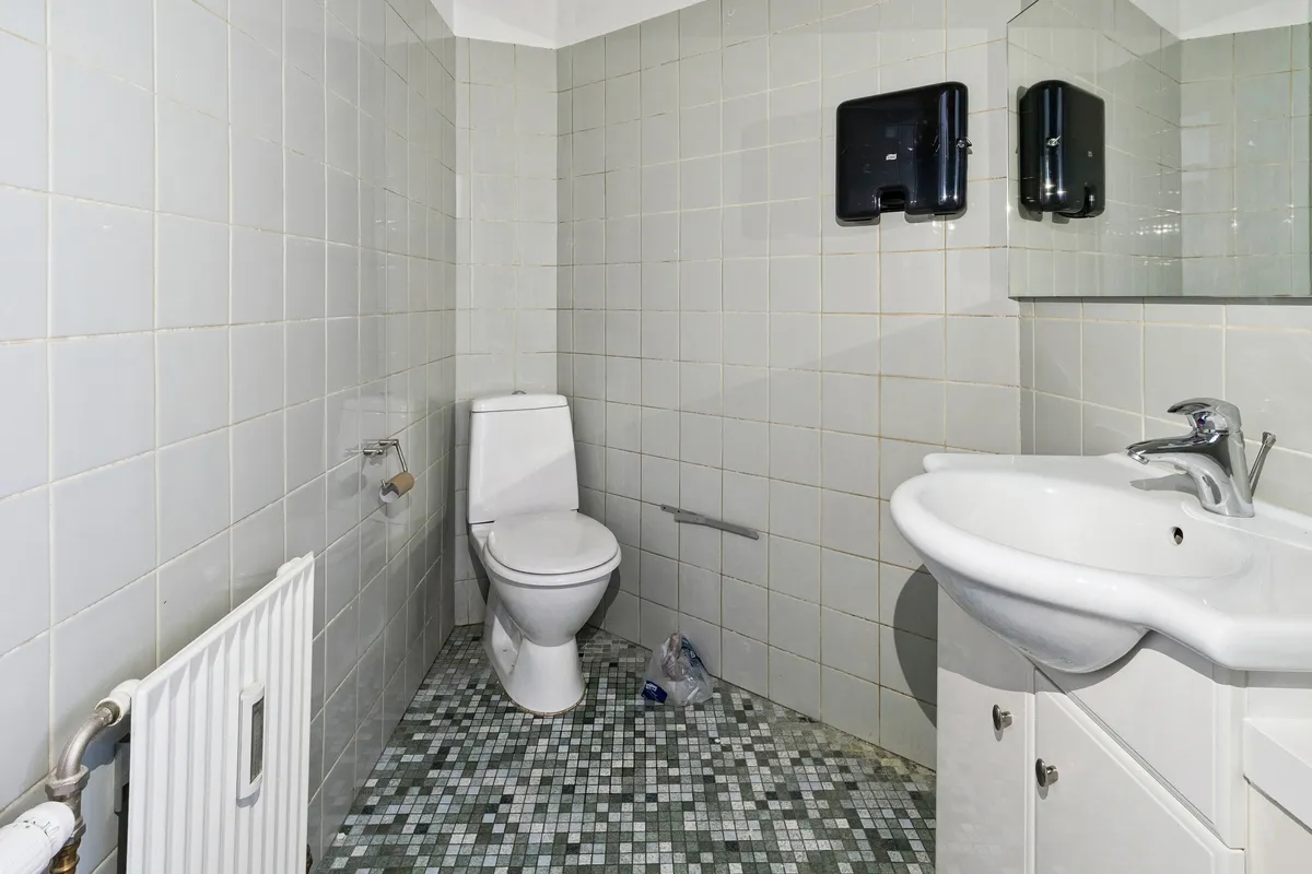 Et lille badeværelse med toilet, håndvask og spejl samt hvide kakeloverflade og radiator, som er installeret på en af væggene.