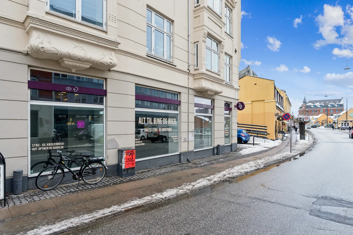 Markant hjørnebutik på Valby Langgade 49B, st. med udsigt over en snelagt vej. Skiltet ALT TIL JONE OG BRER er synligt, og en cykel står parkeret udenfor.
