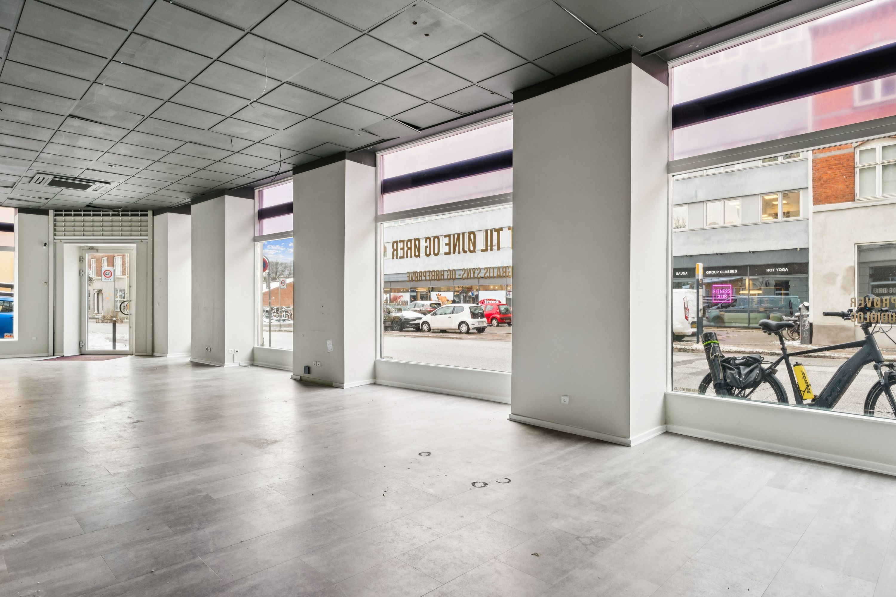 En stor, tom hjørnebutik med store vinduer og lysegule gulve. På gaden er der en bil og en cykel. Butikken er placeret på Valby Langgade.