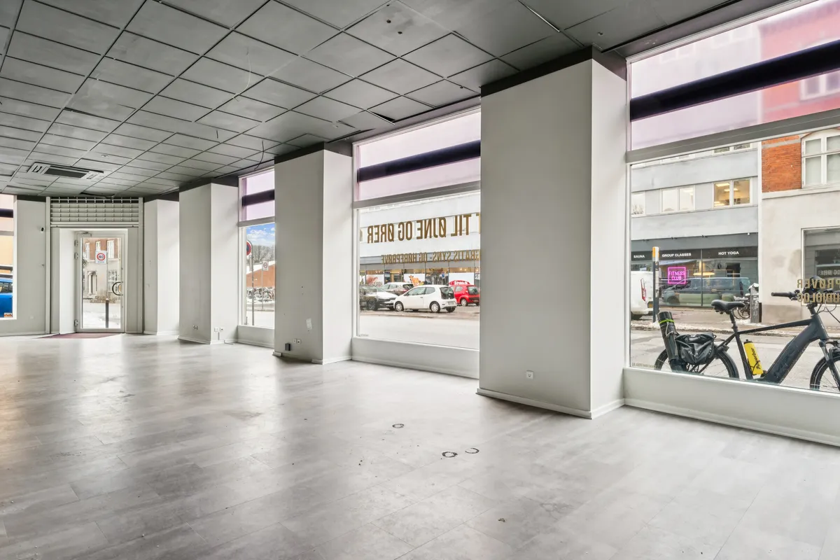 En stor, tom hjørnebutik med store vinduer og lysegule gulve. På gaden er der en bil og en cykel. Butikken er placeret på Valby Langgade.