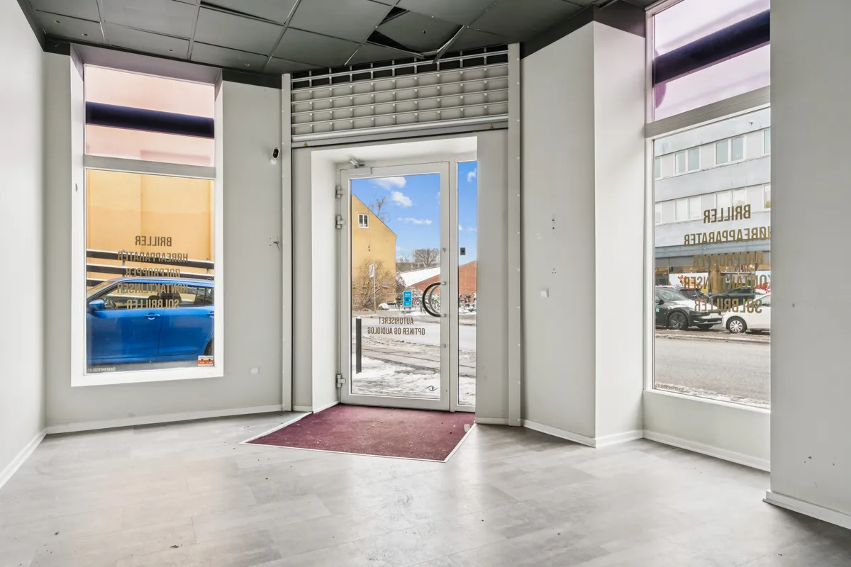 Hjørnebutik på Valby Langgade 49B, st., med glasvinduer og indgangsdør, mærkede skilte og rødt tæppe, udsigt til parkerede biler og bygninger