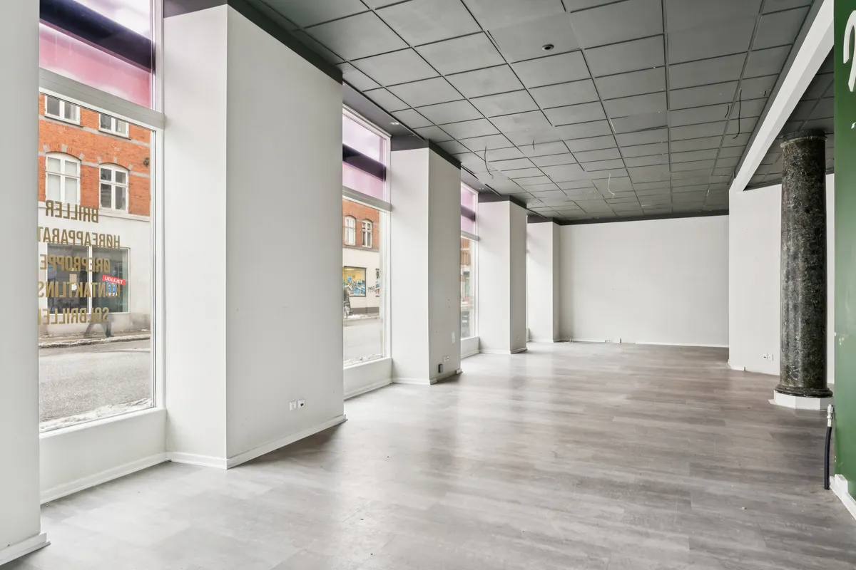 Markant hjørnebutik med store vinduer og lyse lofter, grafiske tekster reflekteret i glas og tømrerede søjler. Tomme gulve med lysegrå belægning og lyse hjørner.