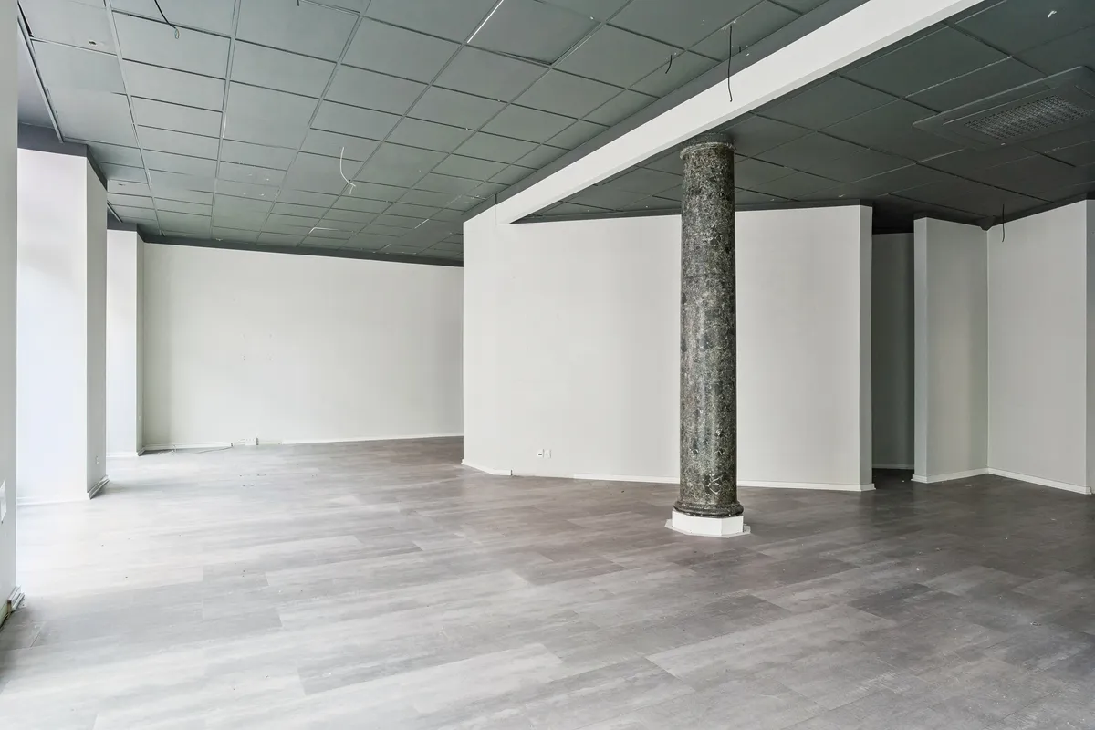 En stor, tom hjørnebutik med højt loft og marmorsøjle, lysegrå gulvbelægning, og hvide vægge på Valby Langgade 49B, st., i området ved Tingstedet.