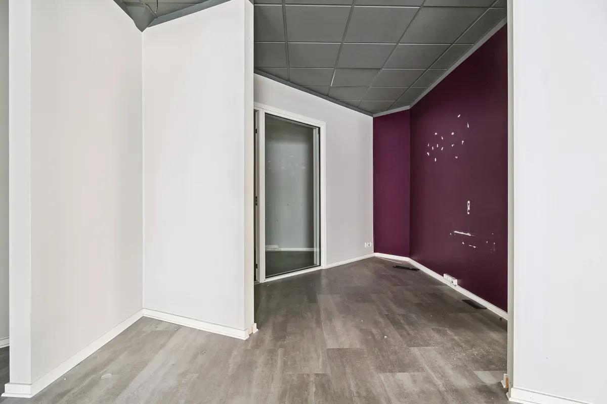 En tom hjørnebutik med hvide og mørke lilla vægge, gulvtæppe og et glasdørsvindue i hjørnet af rummet.