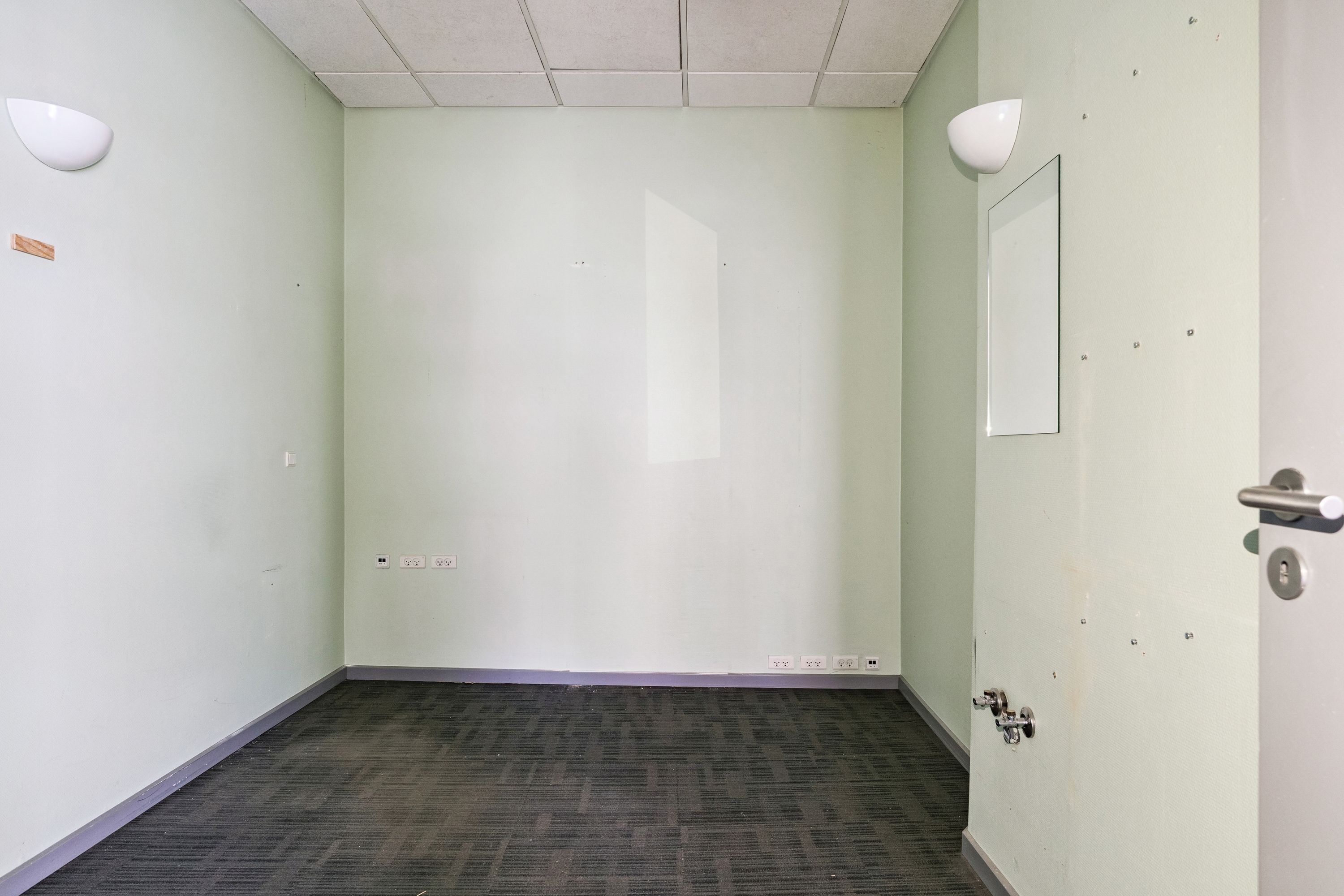 Kort og funktionelt kontor med hvide vægge, grønne loftsplader, hvidt gulv og to hvide væglampe. Der er tre elektriske stikkontakter og et spejl på højre side af døren.