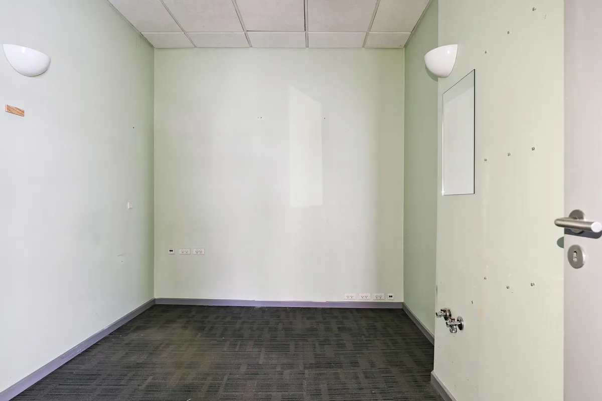 Kort og funktionelt kontor med hvide vægge, grønne loftsplader, hvidt gulv og to hvide væglampe. Der er tre elektriske stikkontakter og et spejl på højre side af døren.