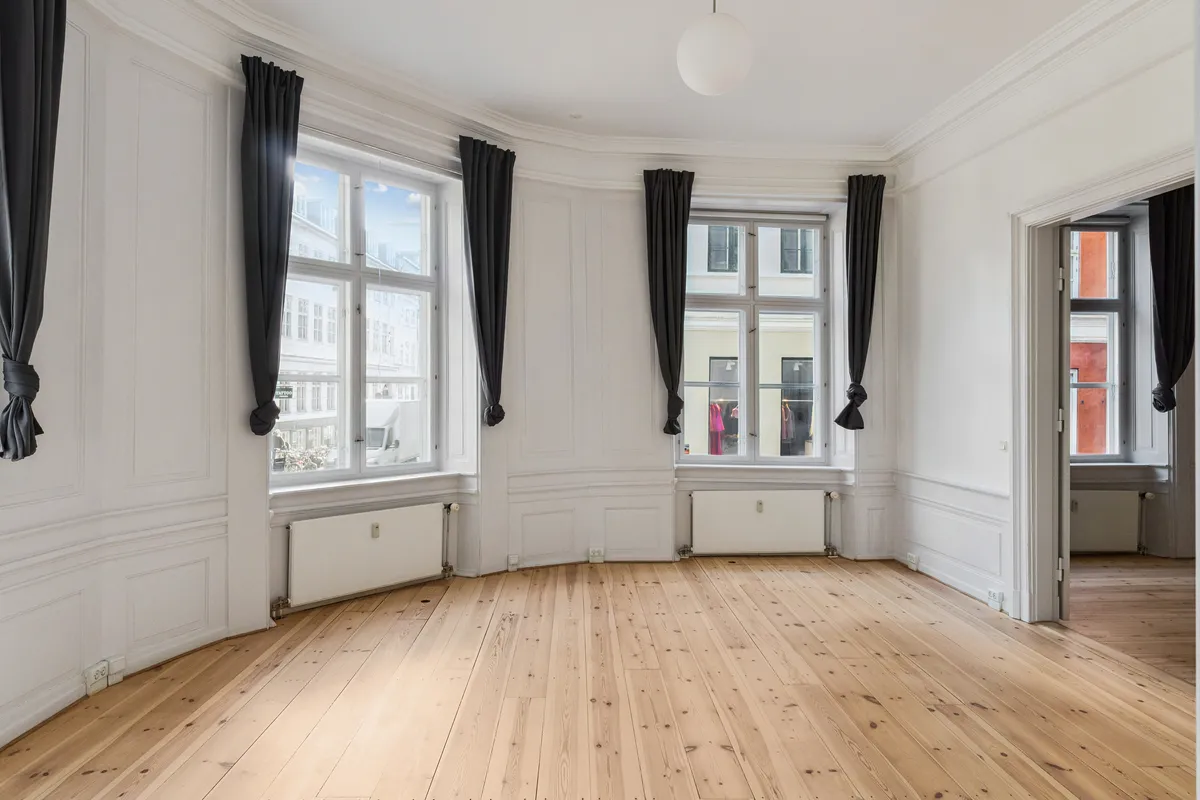 Kontor til leje på Rådhusstræde 6, København. Et lyst og rummeligt lokale med højloft og massiv trægulv. To store vinduer med mørke gardiner.