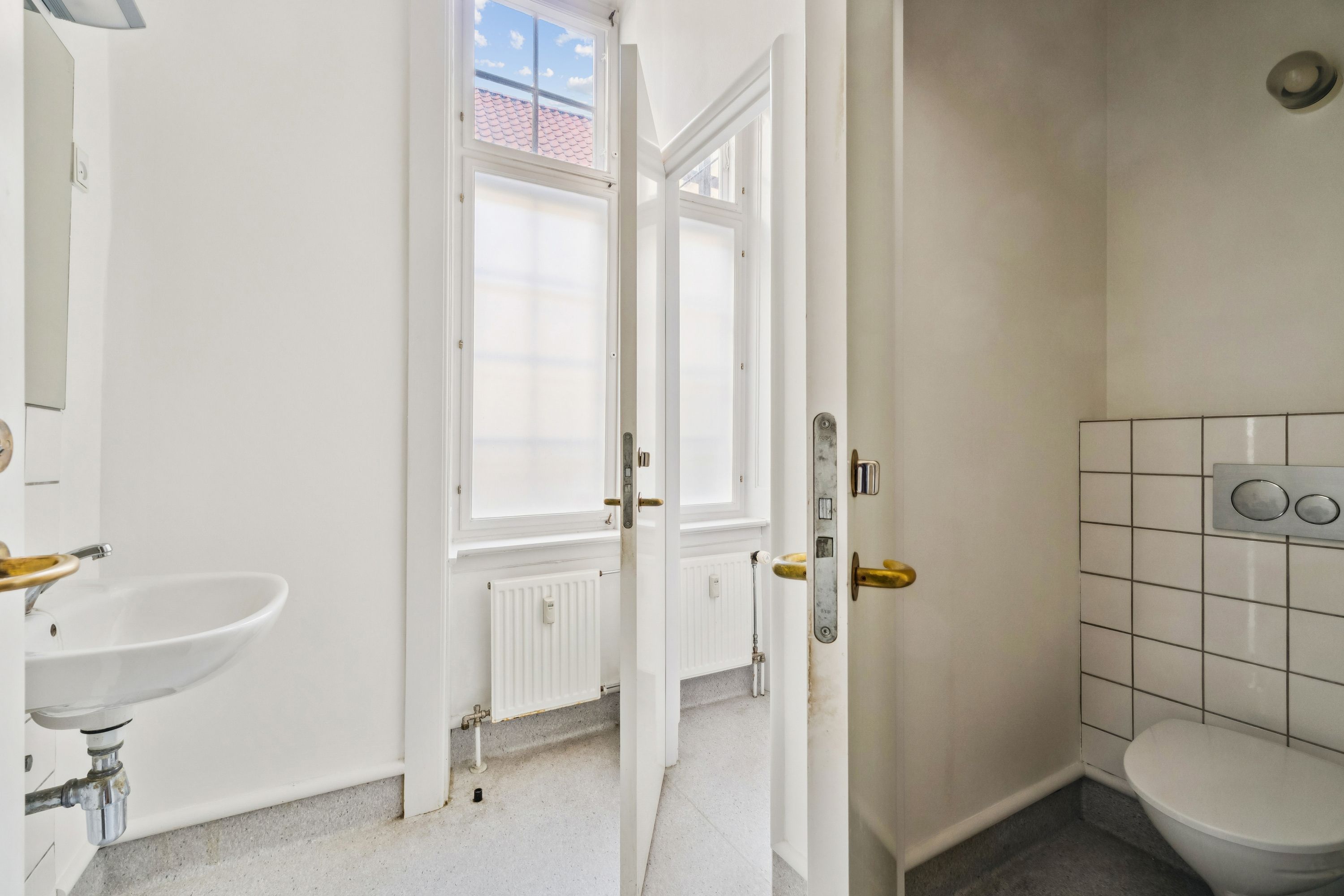 Foto af et rent og moderne badeværelse med en hvid toilet, hvid sænke med en guldfarvet vandhane og et vindue med en udsigt til Rådhusstræde.